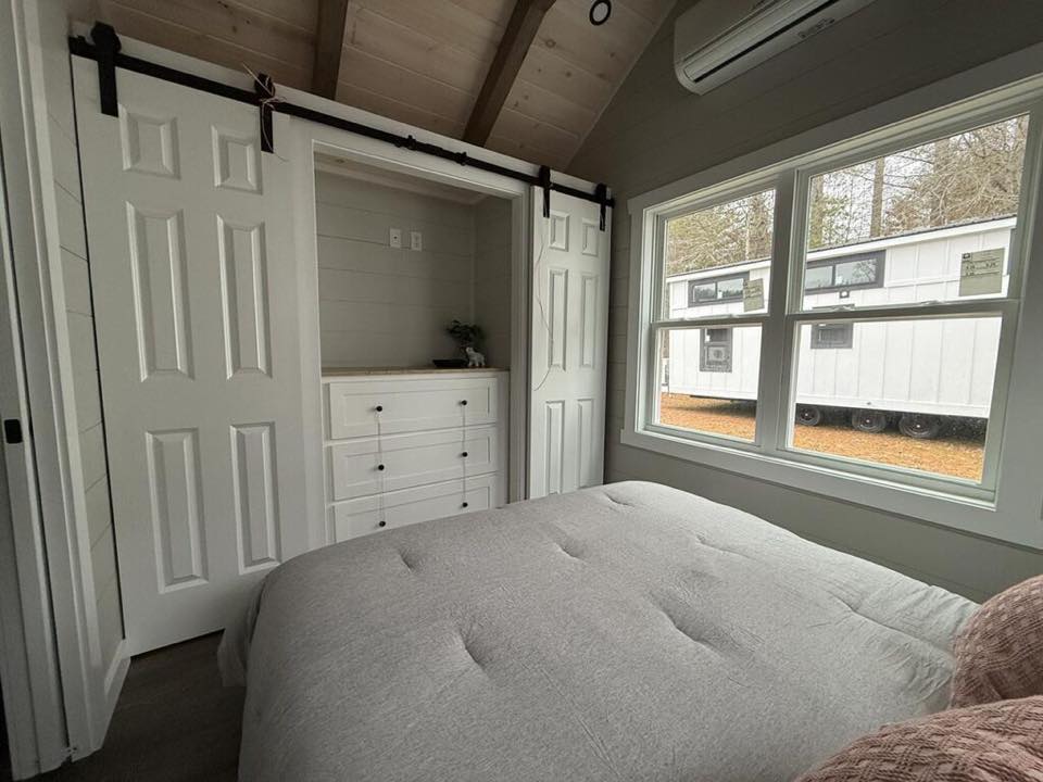 Cozy bedroom with a soft gray bed, white closet with sliding doors, and large window showing a white trailer outside. Warm, peaceful atmosphere.