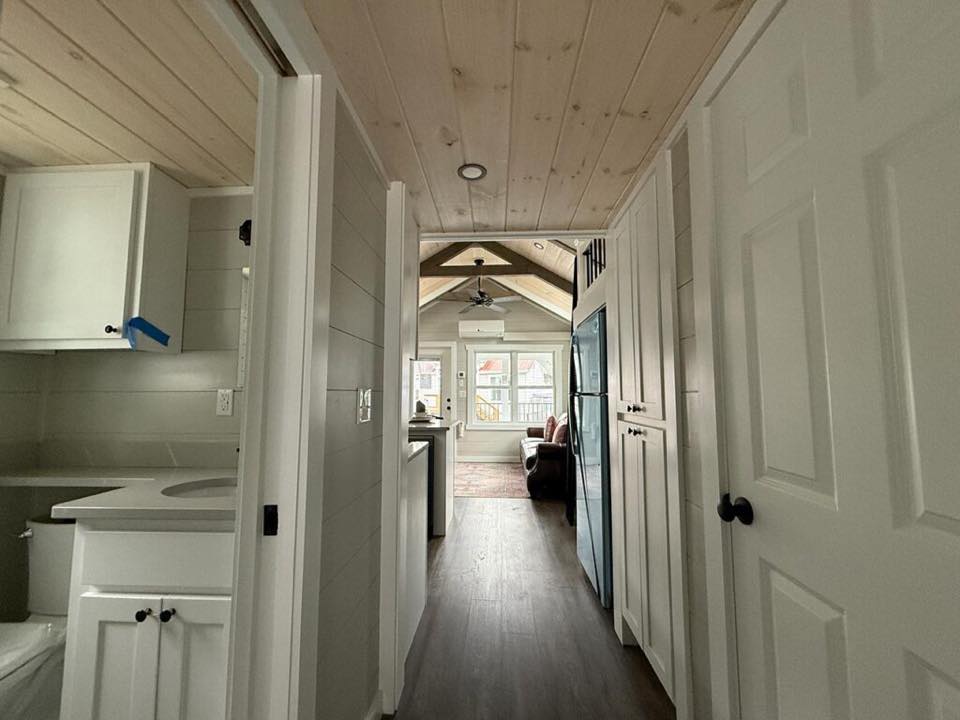 Cozy tiny house interior with wood-paneled ceiling and floor. A narrow hallway leads to a bright living area with large windows and a loft.