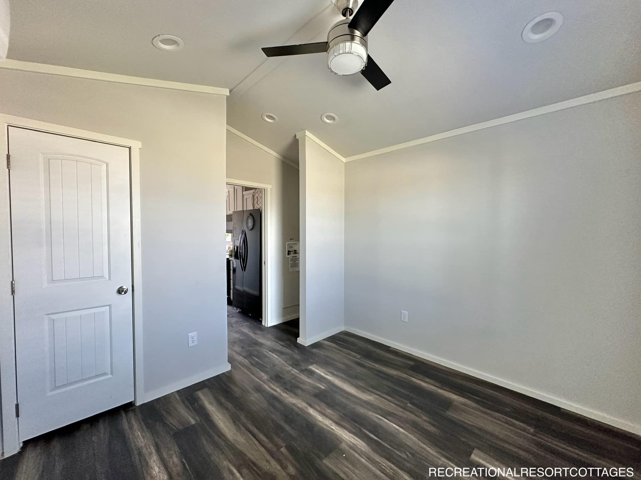 Empty room with light gray walls, dark wood flooring, and a ceiling fan. A doorway leads to a kitchen with stainless steel appliances. Quiet ambiance.
