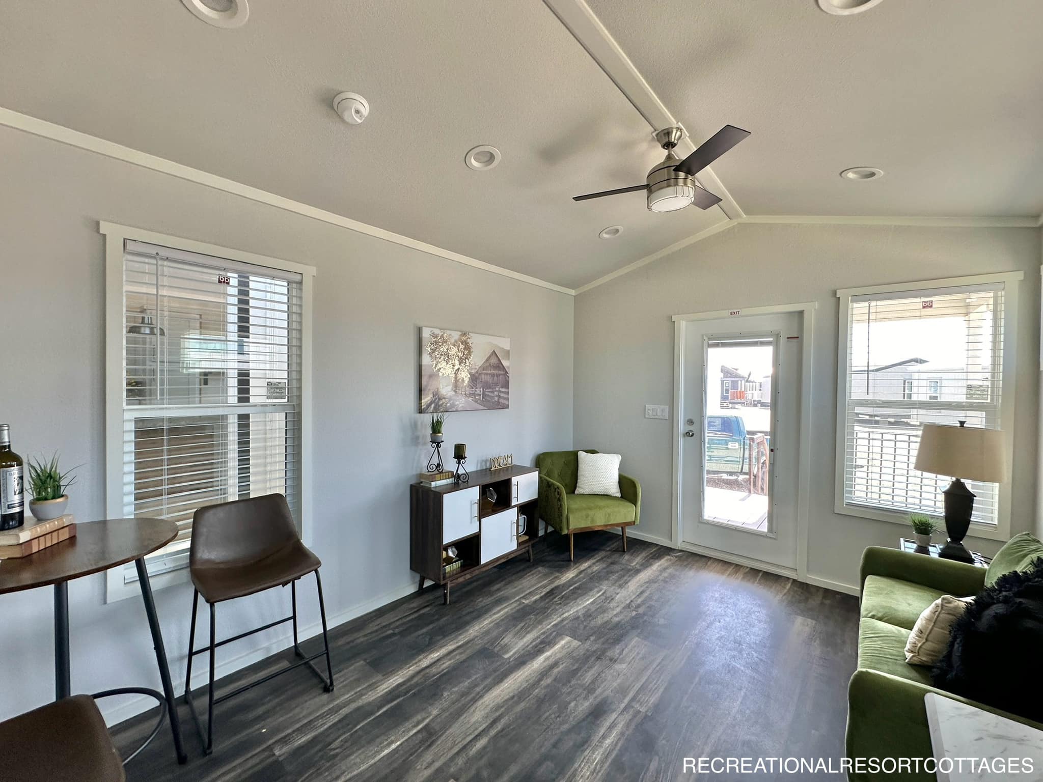 Bright, modern living room with gray walls and dark wood flooring. Features a ceiling fan, green armchairs, and large windows bringing in natural light. A small round table with two chairs and a compact shelf add functionality. Cozy and inviting atmosphere.