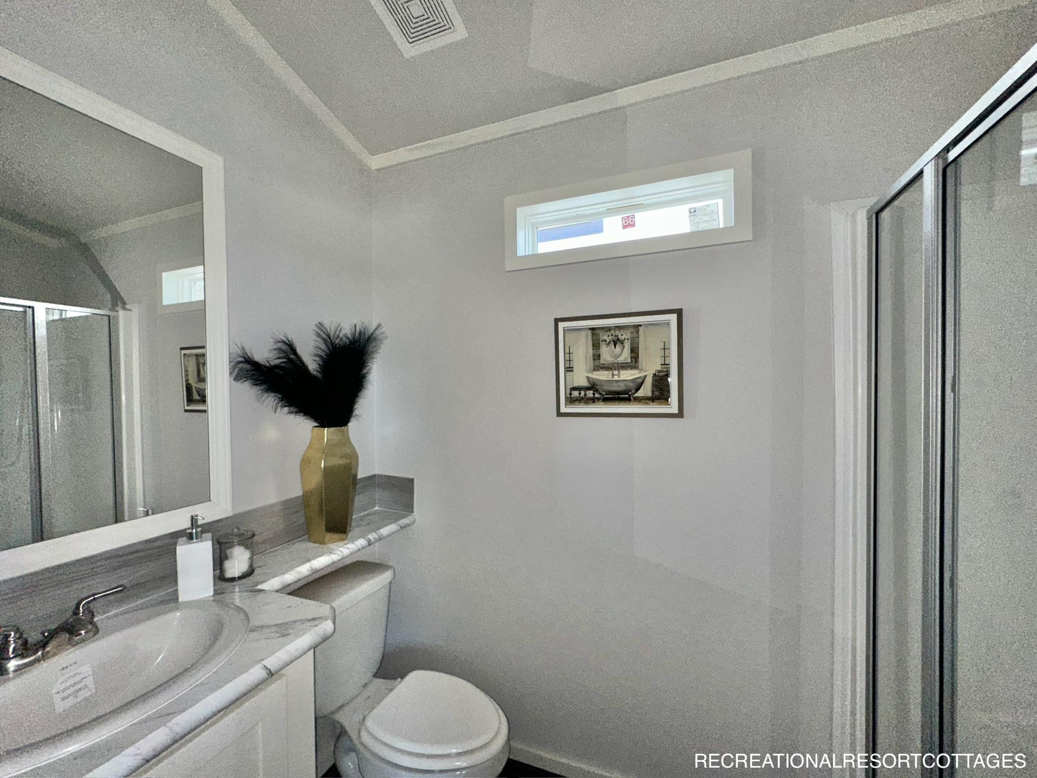 A modern bathroom with a white sink and toilet. A gold vase with black feathers sits on the counter. A small window and framed picture adorn the light gray walls.
