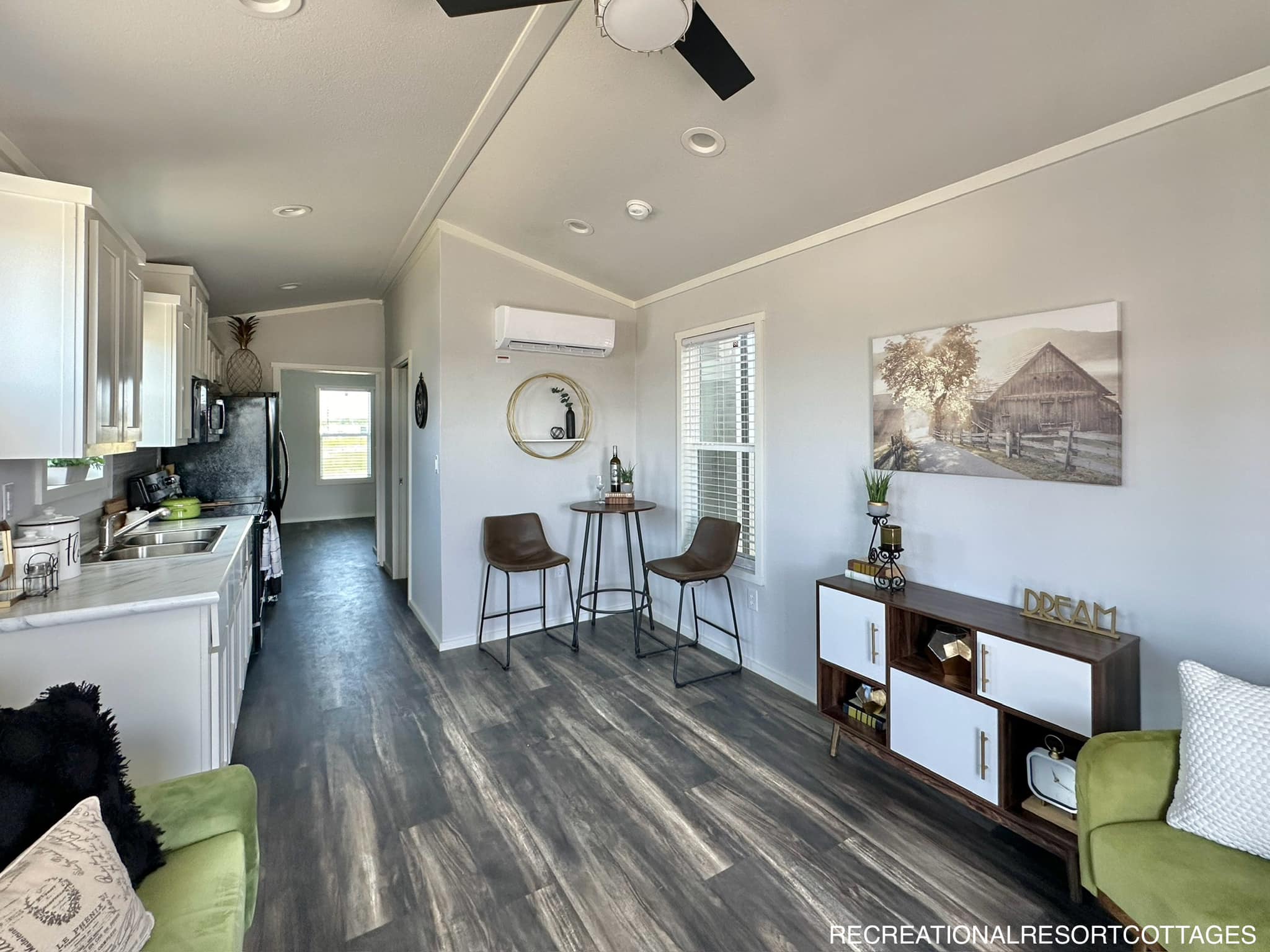 Modern tiny home interior with a cozy kitchen and living area. Features a green couch, bar table with stools, and rustic barn artwork on the wall. Bright and inviting.