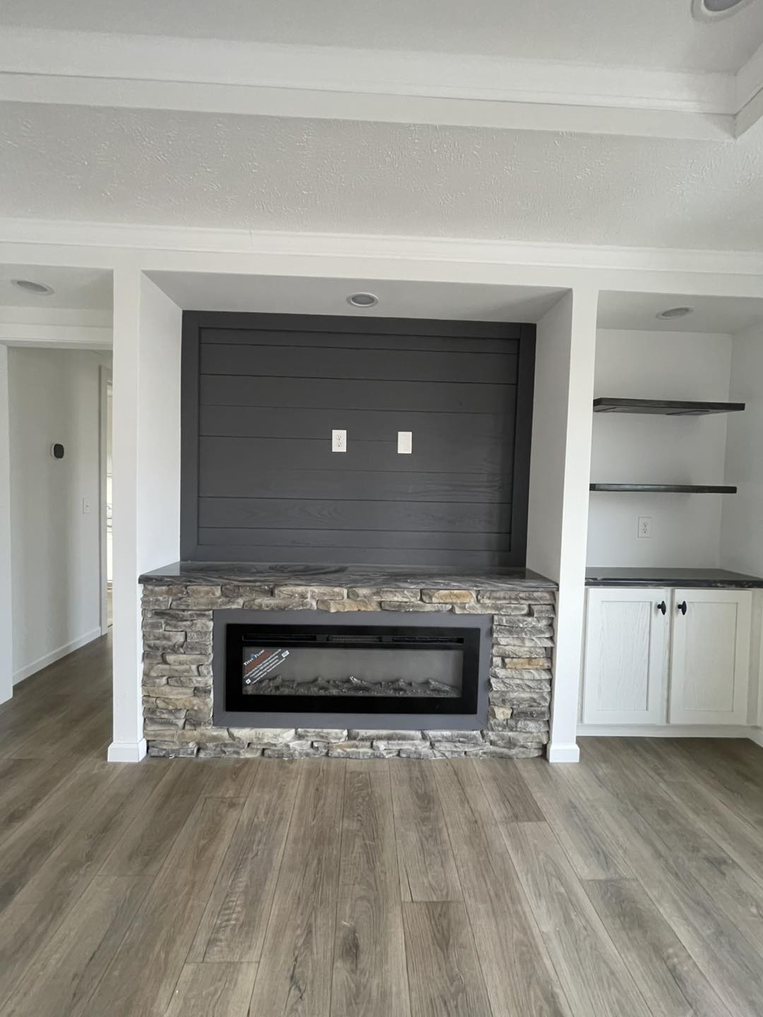 Modern living room with a stone-framed fireplace set in a white wall. Dark shiplap paneling above, flanked by shelves and white cabinets, all on wood flooring.