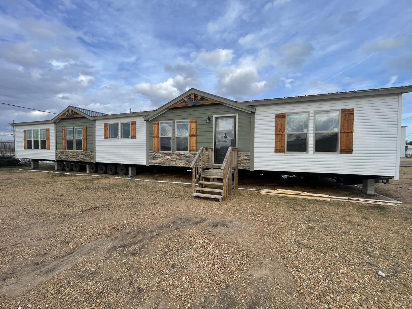 Large manufactured home with a gabled roof, wooden shutters, and stone accents sits on gravel under a partly cloudy sky, conveying a rustic charm.