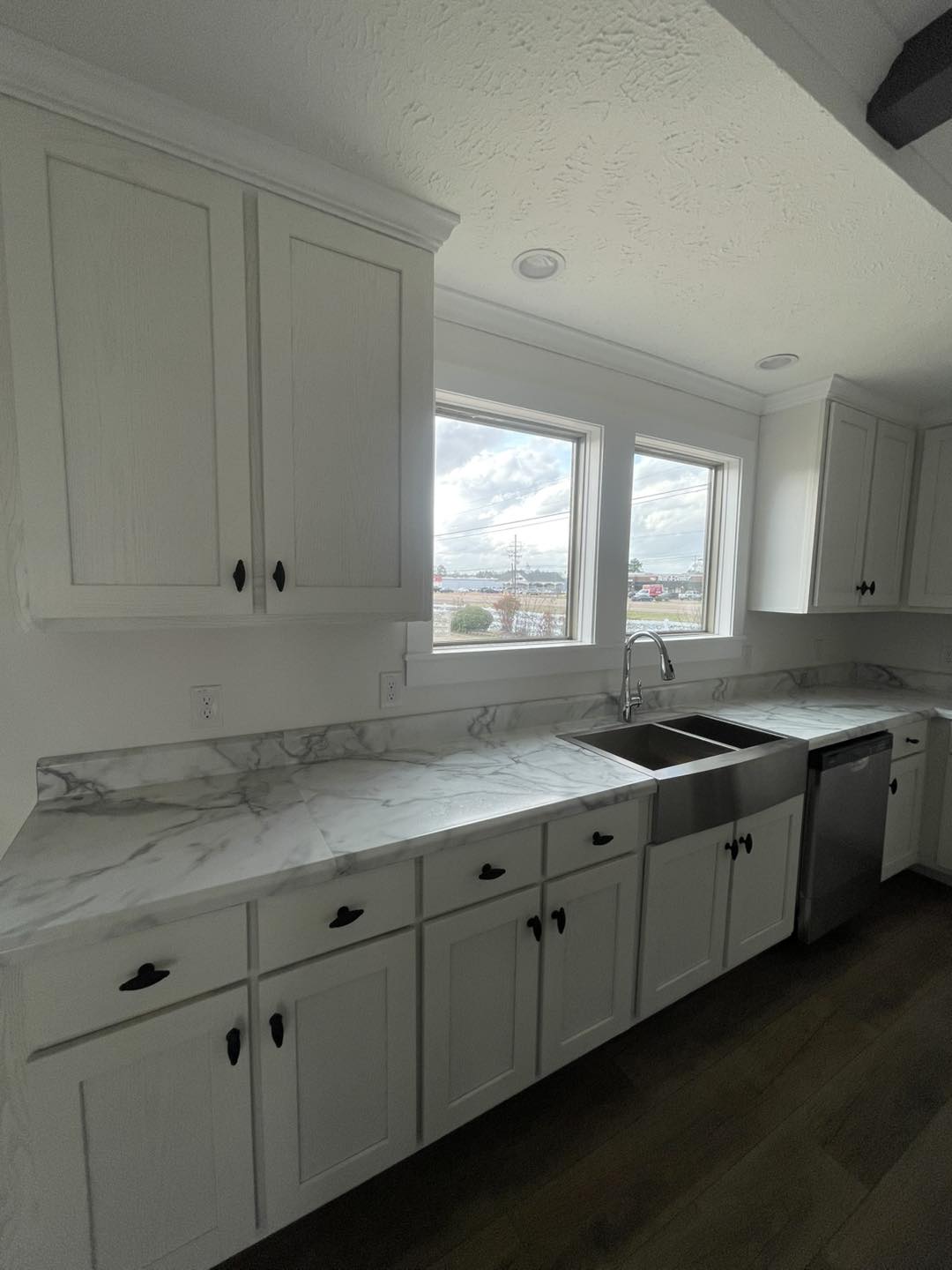 Bright kitchen with white cabinets and marble countertops under a large window. Includes a stainless steel sink and dishwasher, creating a clean, modern look.