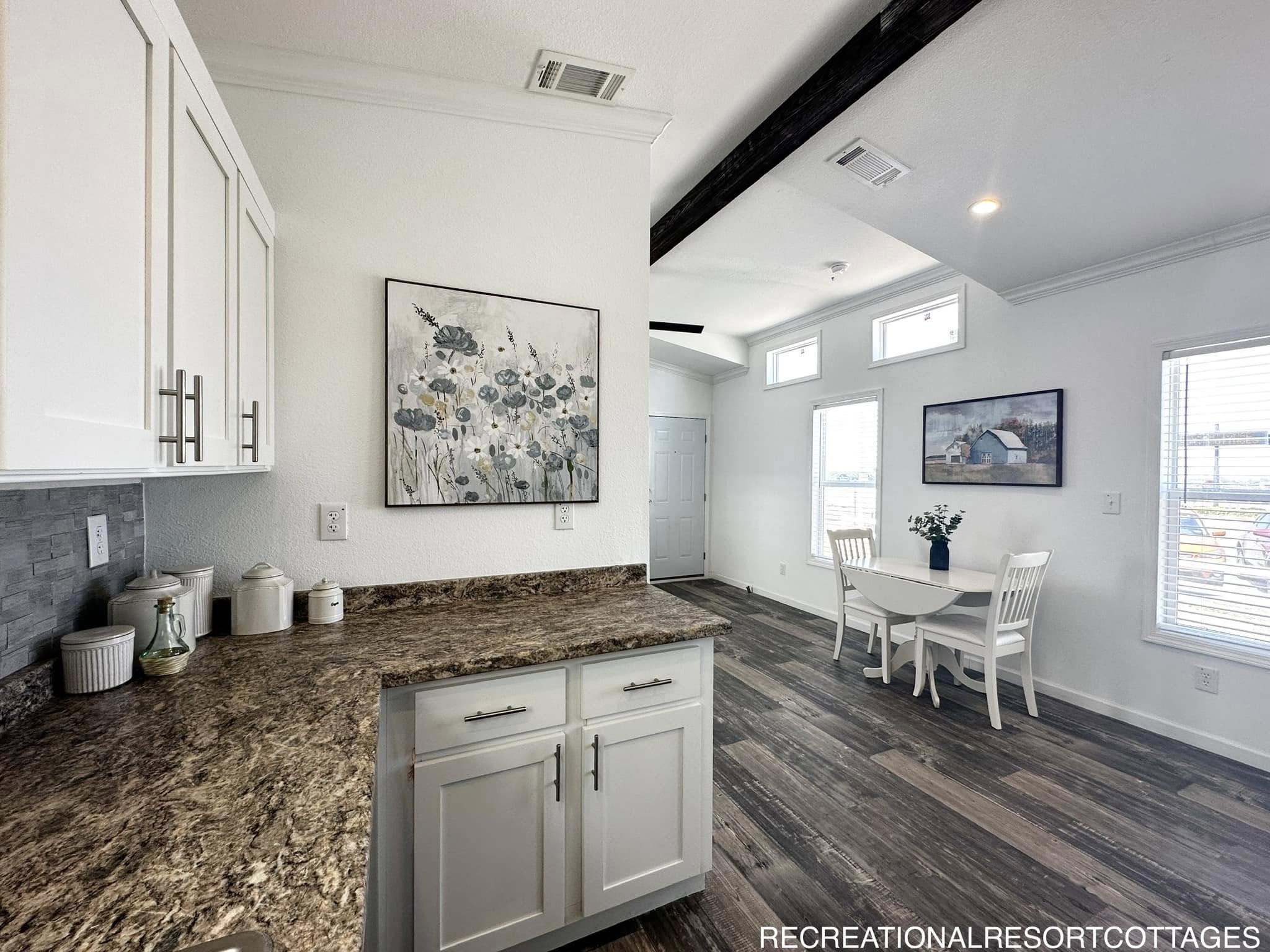 Modern kitchen and dining area with white cabinets and a gray countertop. A floral painting hangs above the counter. Wood floors lead to a round table set by large windows, creating a bright, airy space.