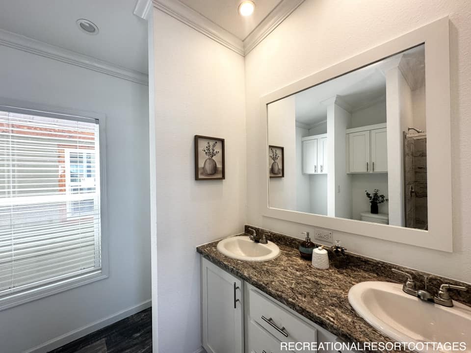A bright bathroom features a granite countertop with dual sinks, white cabinetry, and a large wall mirror. A window with blinds allows natural light.