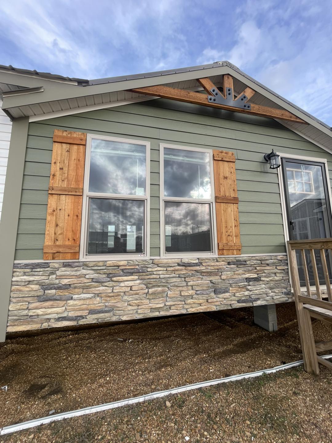 House exterior with green siding and wooden accents, featuring large windows with wood shutters, stone veneer, and a small porch. Cloudy sky above.