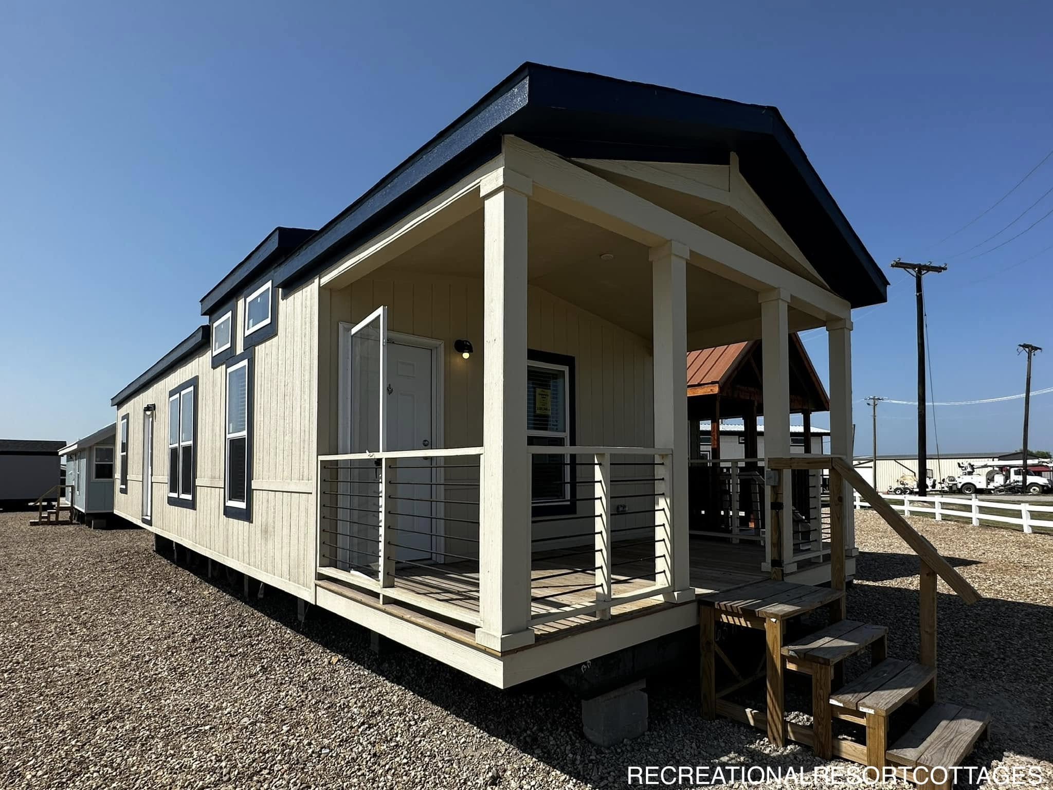 Alt text: A beige modular home with a dark blue roof is situated on a gravel lot under a clear blue sky. The home features a small wooden porch with railings and steps leading up to the entrance. In the background, there are other similar homes and utility poles. The atmosphere appears calm and quiet.