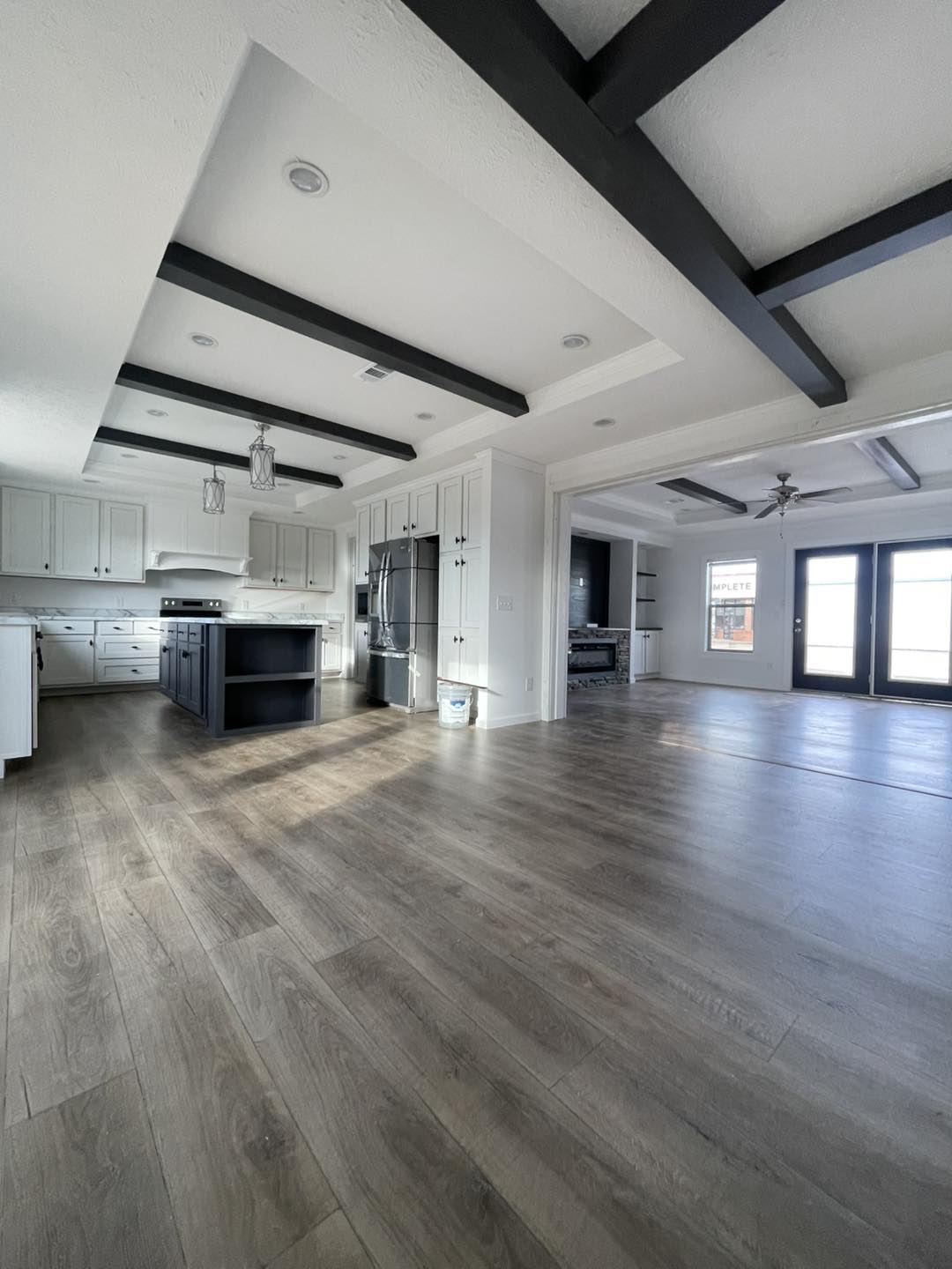 Spacious modern kitchen with light wood floors, black-beamed ceiling, white cabinets, and central island. Open layout flows into a sunlit living room.