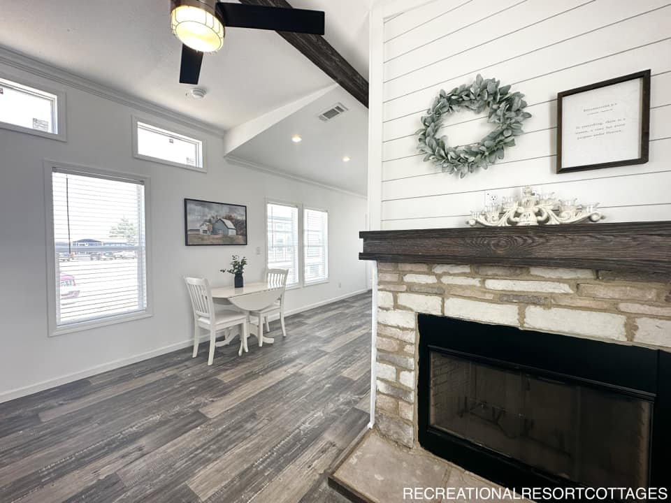 Charming living area with rustic fireplace, shiplap walls, and wreath decor. Nearby, a cozy dining table with white chairs, beneath large windows. Cozy and inviting.