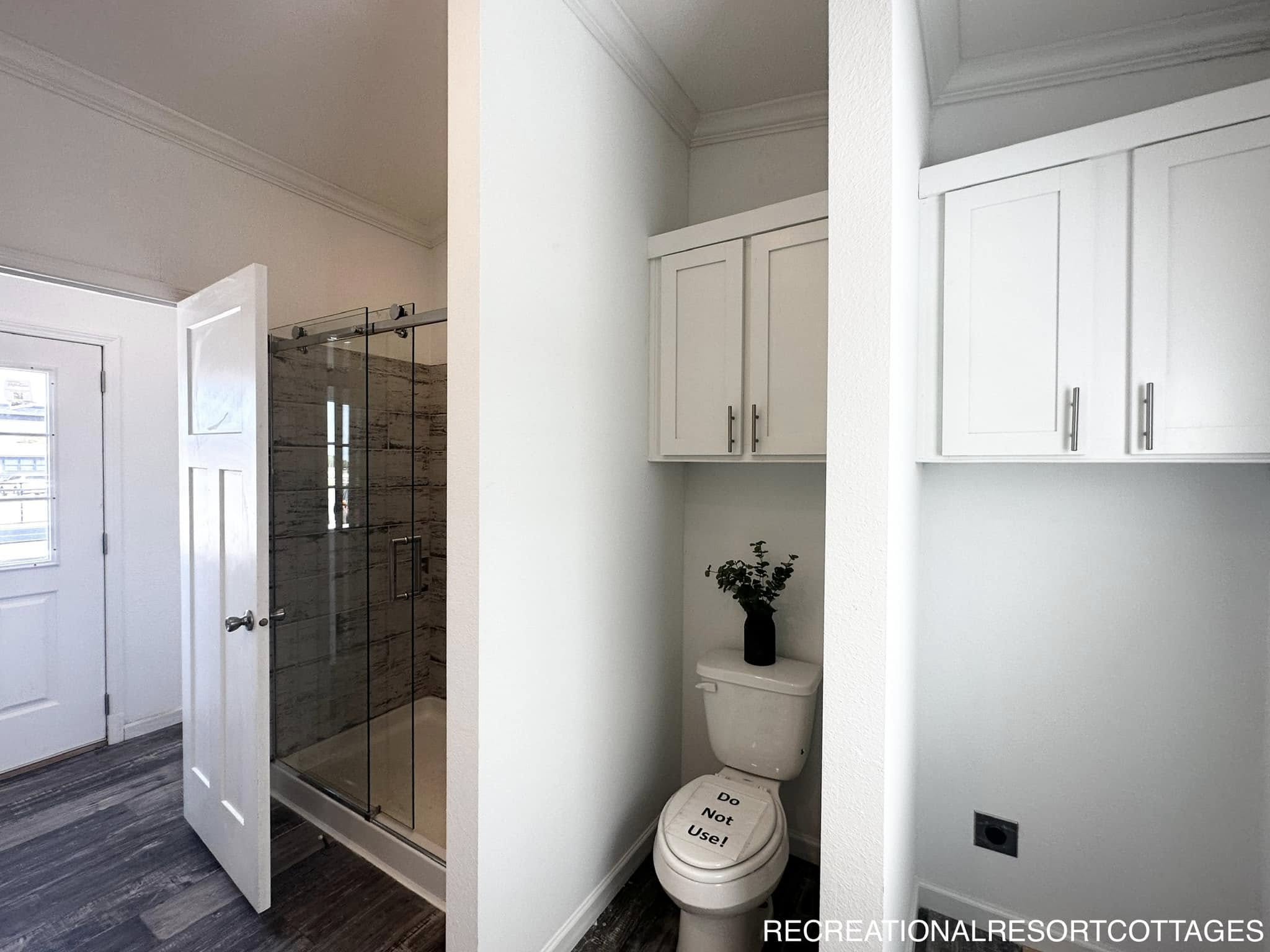 Modern bathroom with glass shower door, white cabinetry above a toilet, and a small plant. A sign on the toilet reads "Do Not Use!"