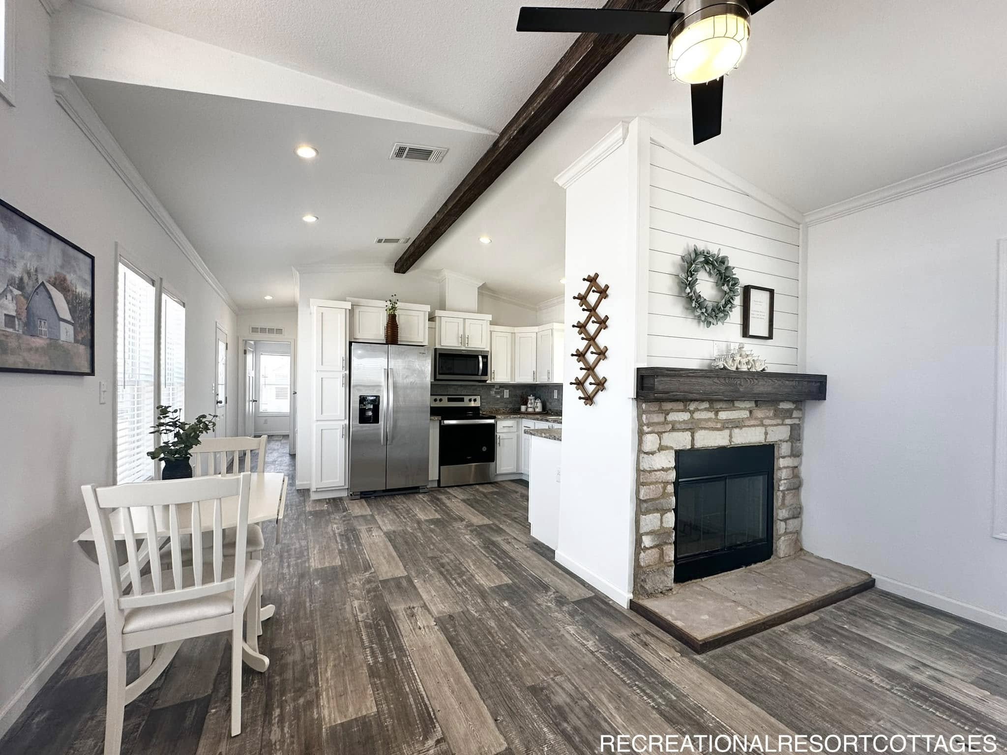 Bright, modern kitchen and dining area with wood flooring. Features white cabinets, stainless steel appliances, and a cozy stone fireplace with a wreath.
