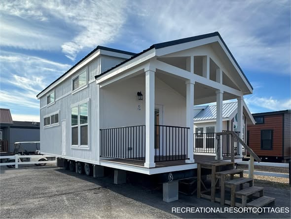 A modern white tiny house on wheels with a peaked roof, small porch with railings, and steps leading up. Clear sky, other homes in the background.