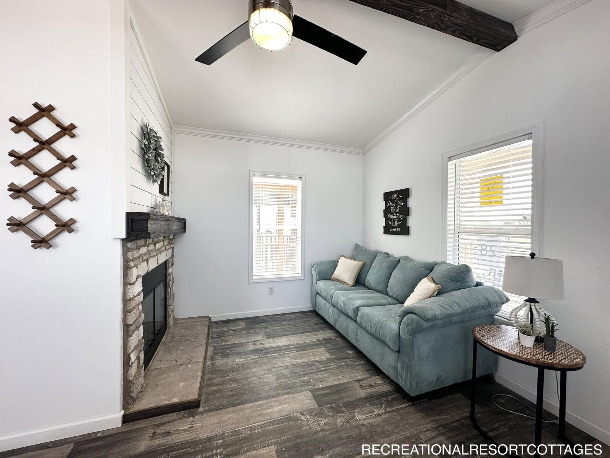 Cozy living room with stone fireplace, teal sofa, throw pillows, wooden side table with lamp, wood flooring, black ceiling fan, and wall decor.