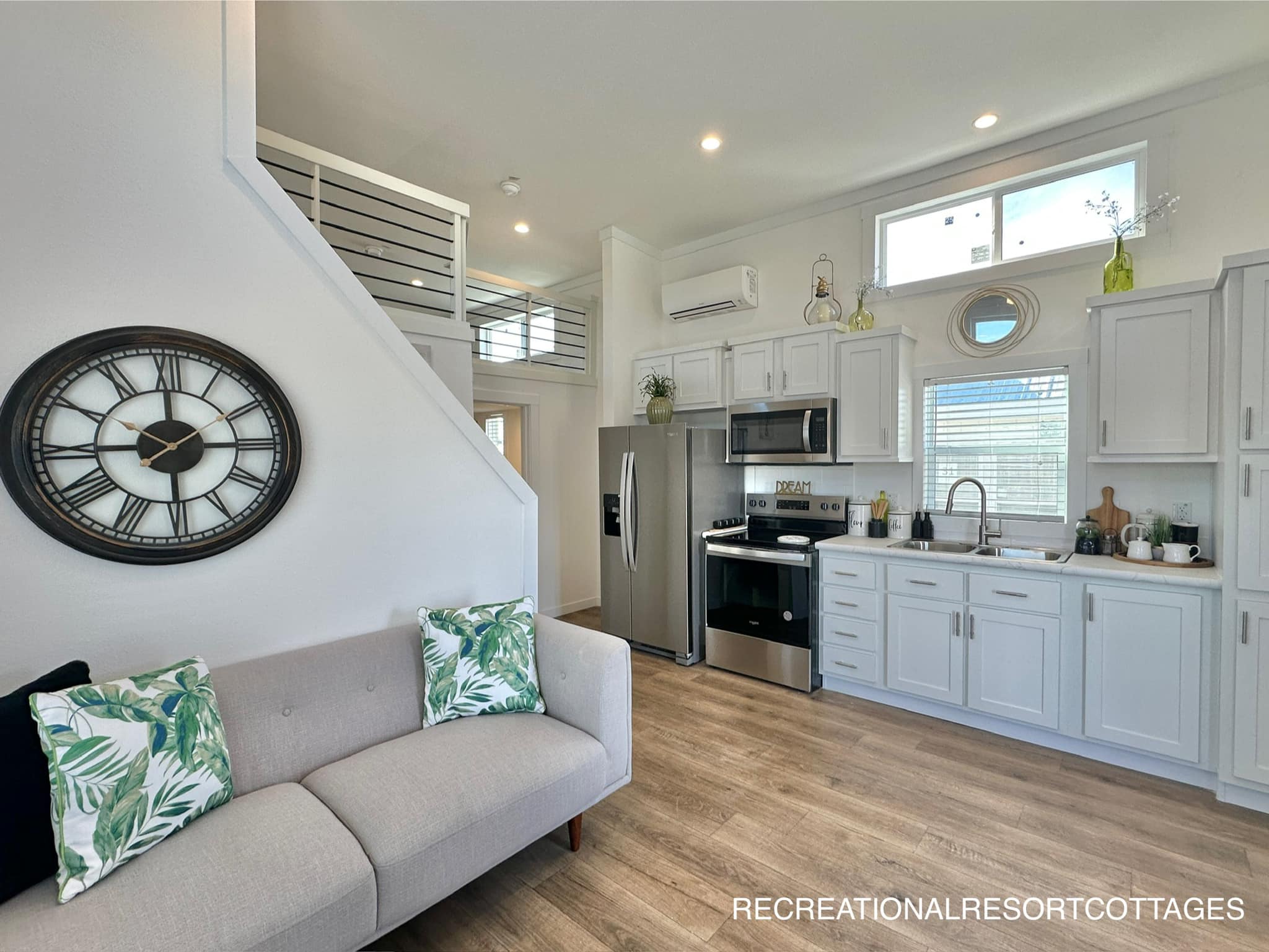 Modern tiny home interior with a cozy gray sofa, tropical pillows, and a large clock. Bright kitchen features white cabinets and stainless steel appliances.