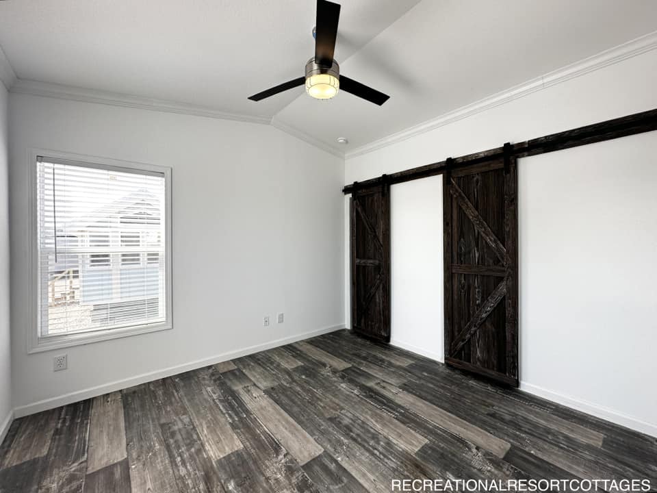 Empty room with white walls and dark wood flooring, featuring two rustic barn doors and a ceiling fan. Light from a window with blinds creates a bright, airy atmosphere.