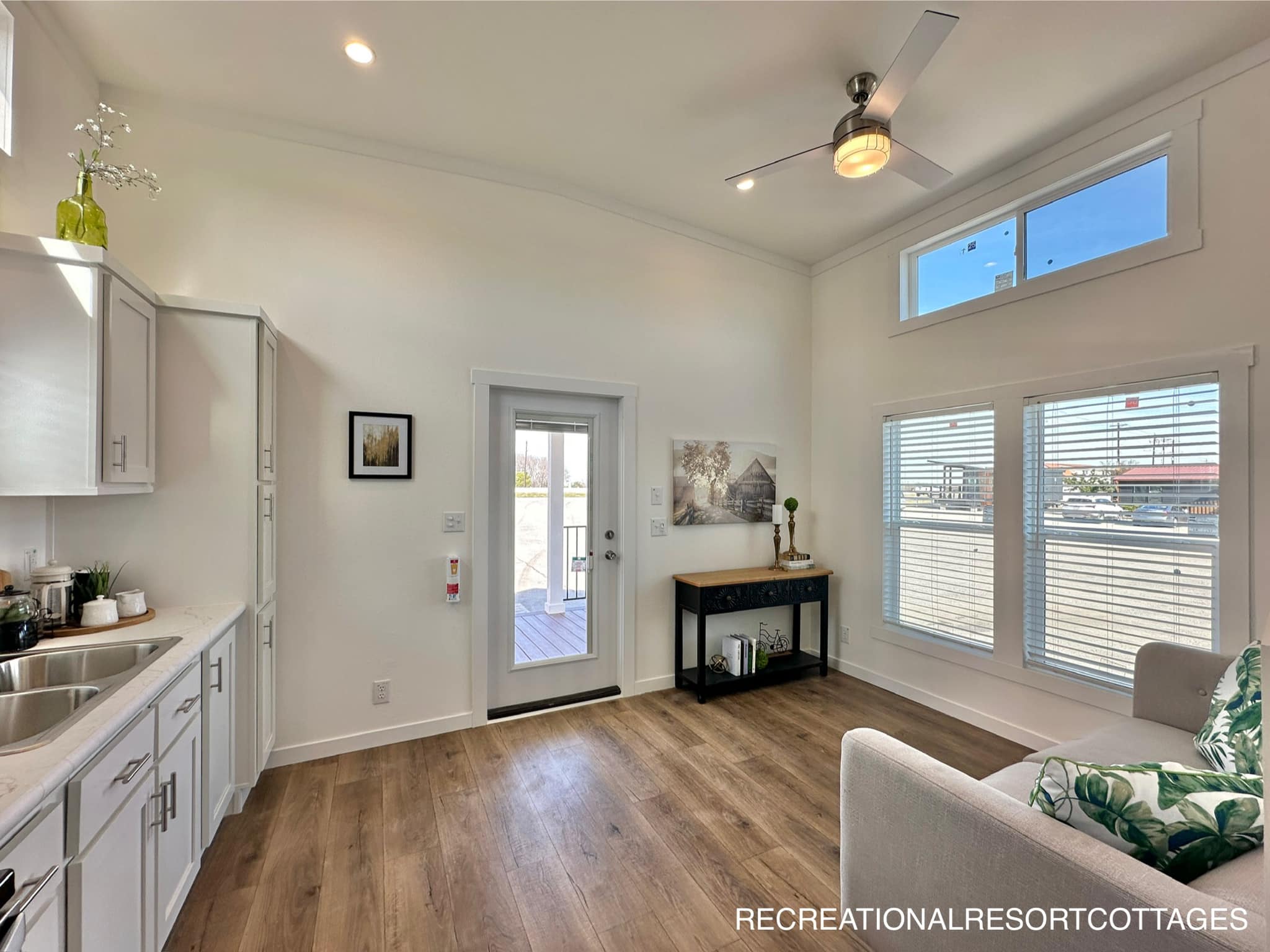 Bright, modern living area with white walls, hardwood floors, and ceiling fan. Large windows and glass door let in natural light. Minimalist decor.