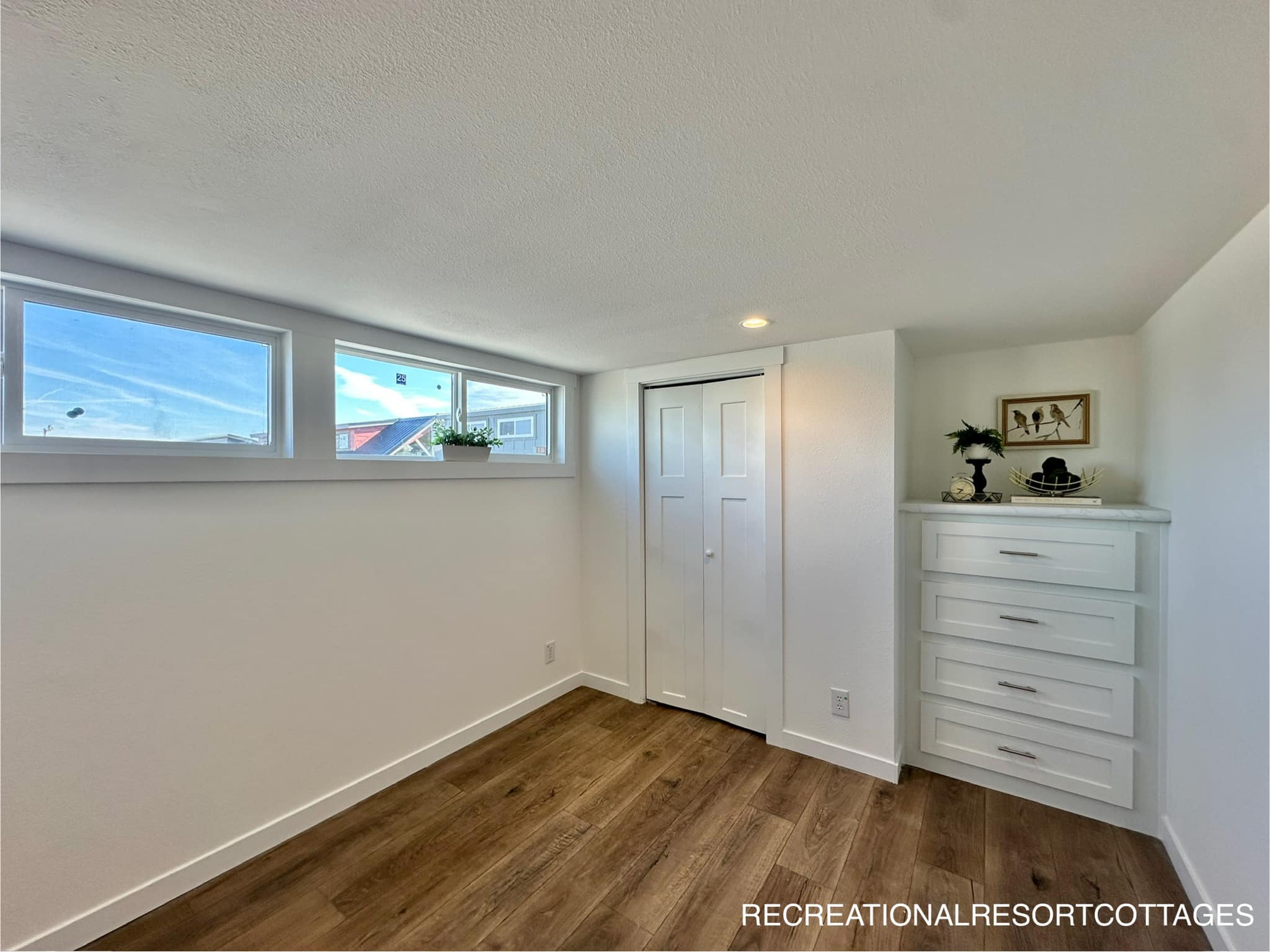 Bright room with wood flooring, featuring narrow horizontal windows, a small closet with white double doors, and a chest of drawers with decor on top.