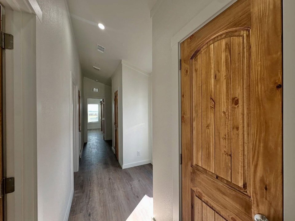 A narrow, well-lit hallway with light wooden floors and white walls. There are several wooden doors along the hallway, leading to a sunlit area at the end.