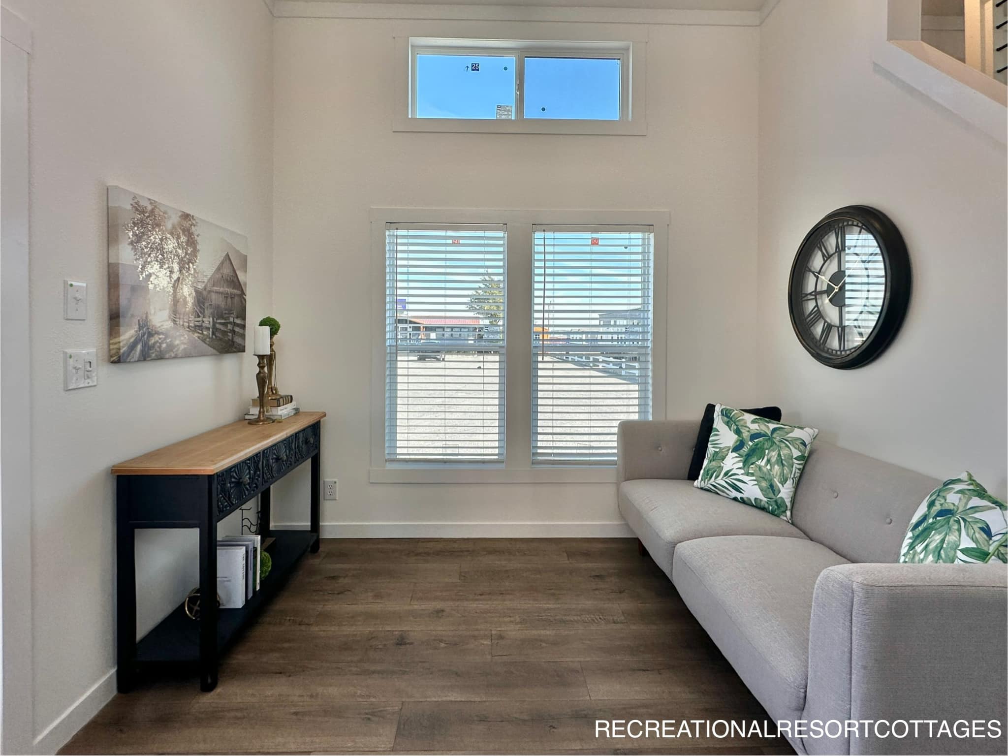 A cozy living room with a large window and blinds. The gray sofa has tropical-patterned cushions. A black clock is on the wall, and a wooden console table with decor is on the left.