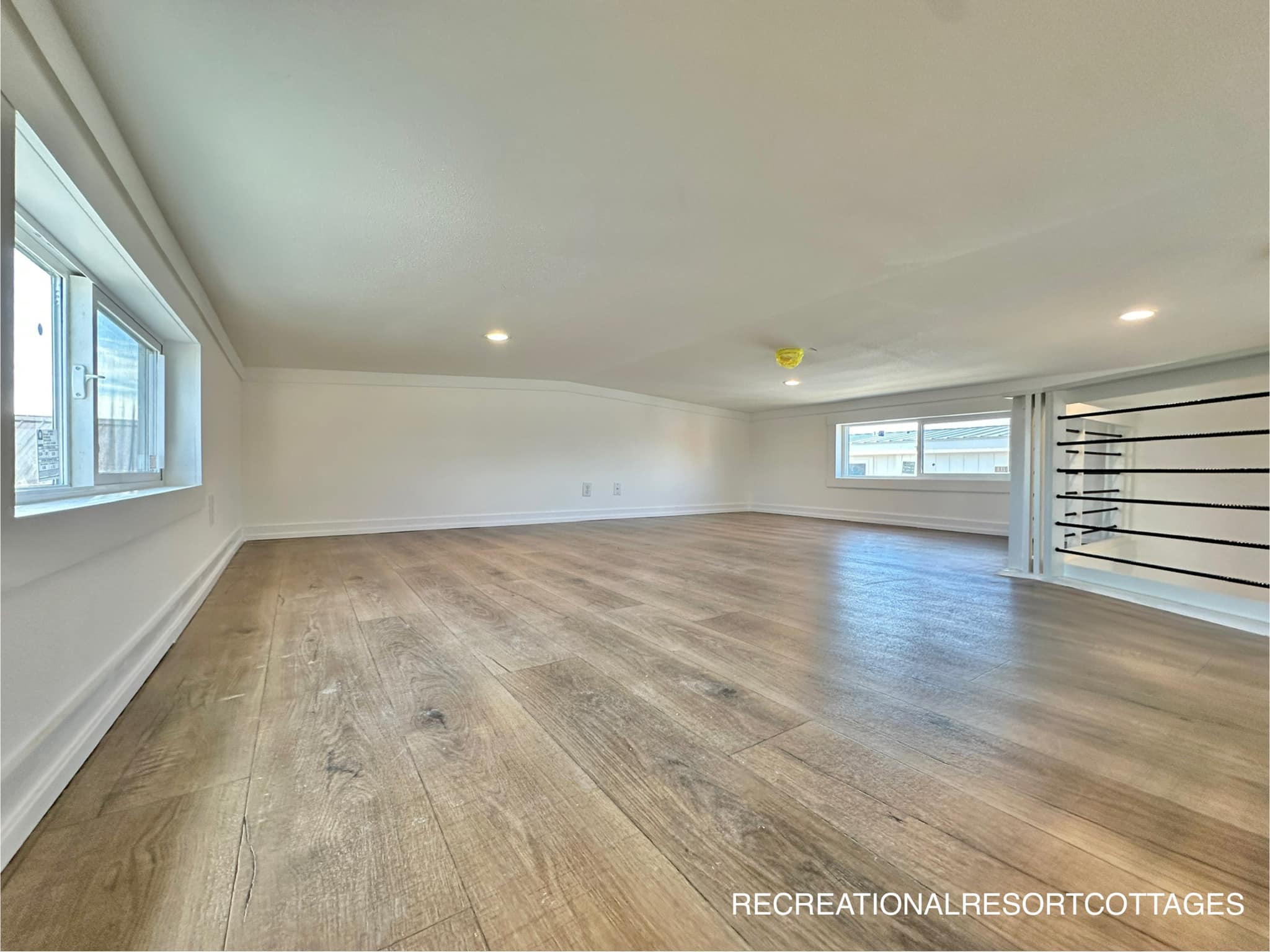 Spacious room with light wood flooring and white walls under a low ceiling. Large windows on the left and back provide natural light. Calm, airy atmosphere.