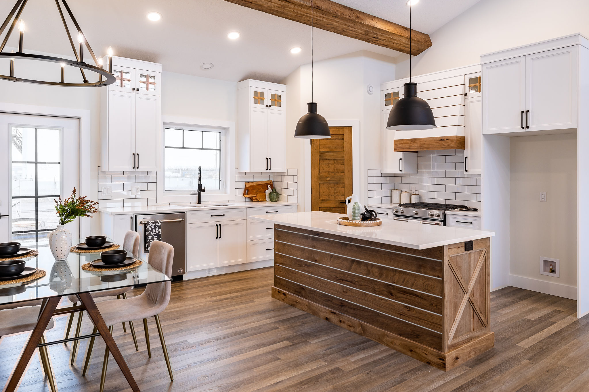 Modern kitchen with white cabinets, a wood island, and pendant lights. A glass dining table with black settings adds elegance. Bright and spacious.
