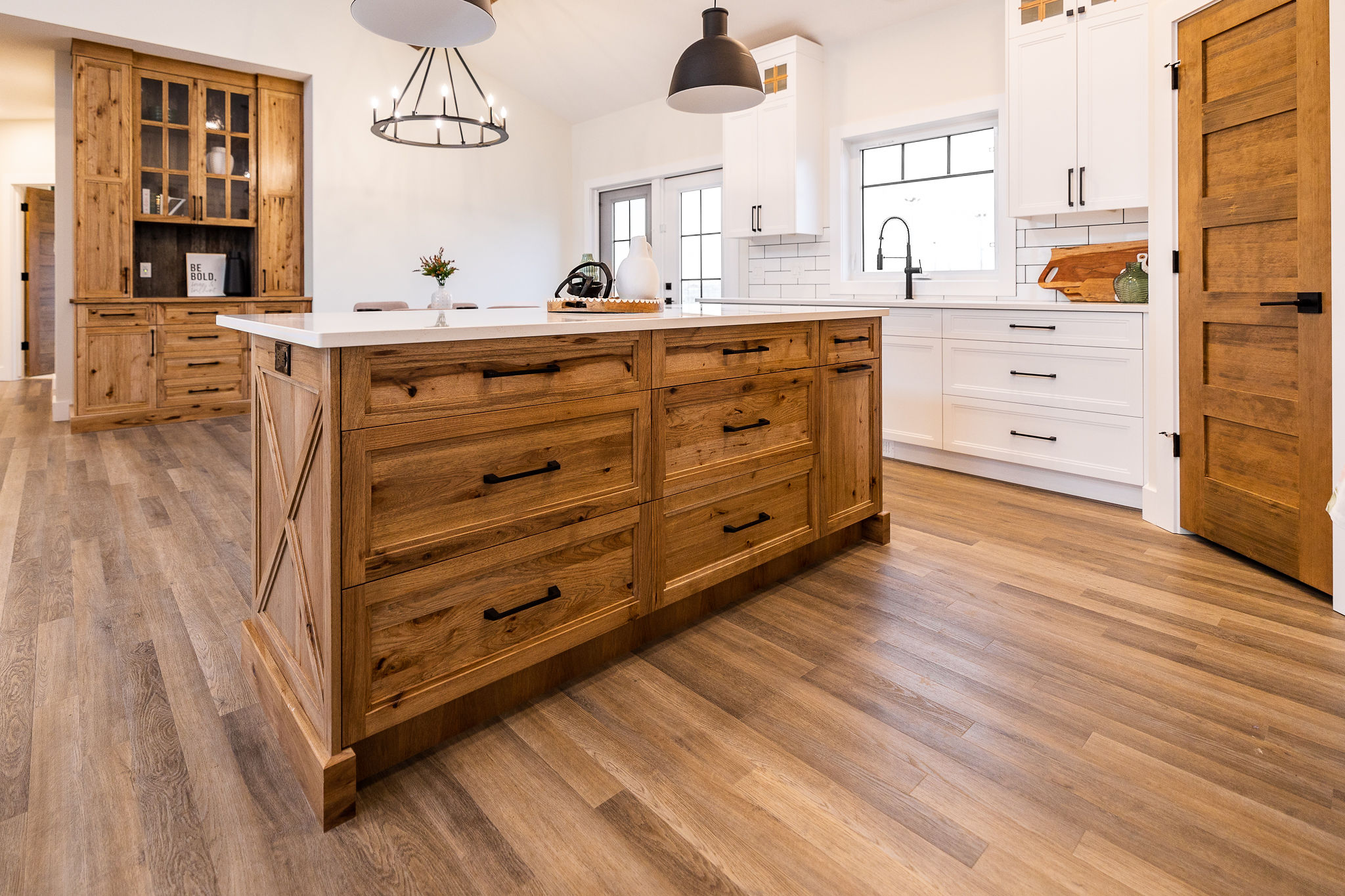Modern kitchen with a rustic feel, featuring a large wooden island, pendant lights, white cabinets, and wooden flooring. Bright, warm ambiance.