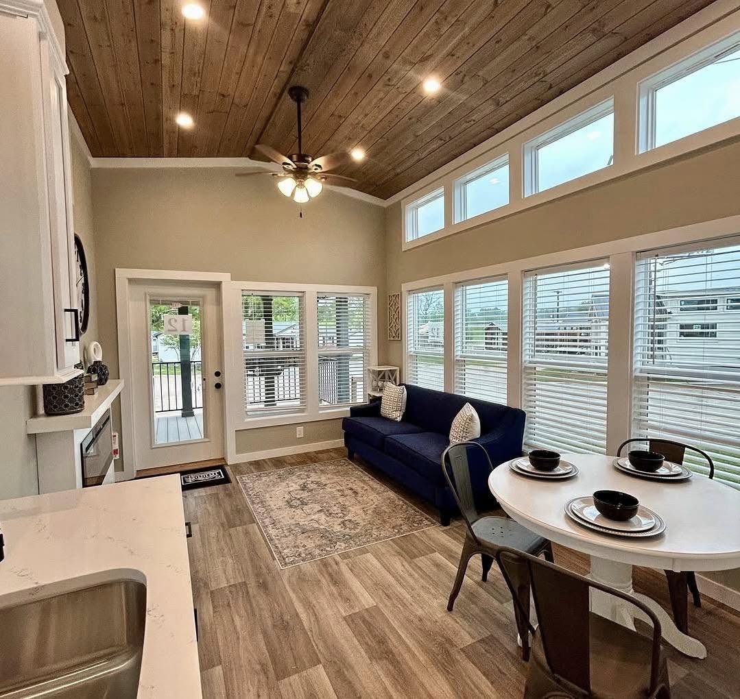 Stylish living area with a wooden ceiling, ceiling fan, large windows with blinds, blue sofa, dining table with dishware, and wood flooring. Cozy and bright.