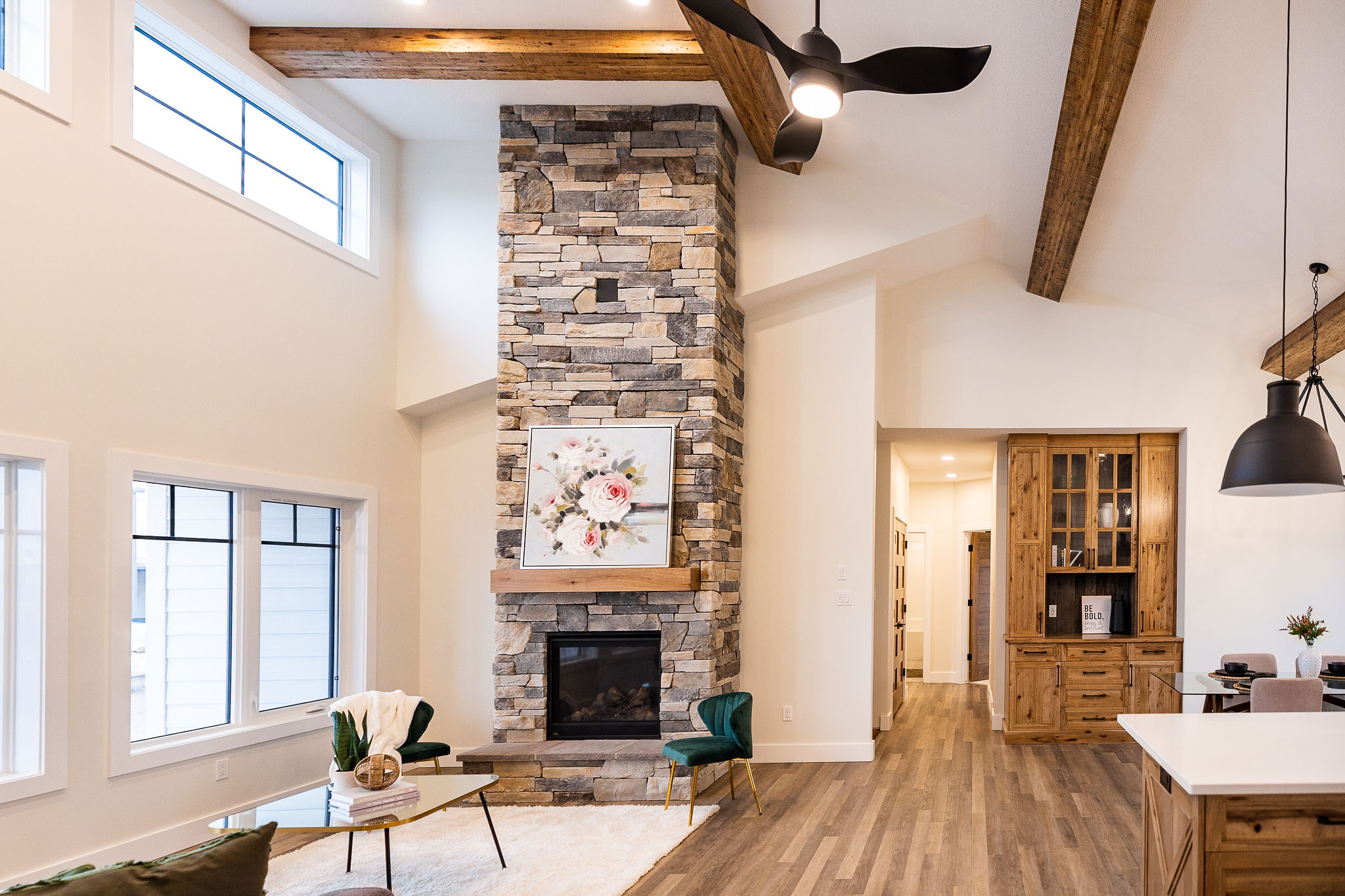 Spacious living room with a stone fireplace, floral artwork above it. Wooden beams on the ceiling. Modern kitchen visible to the right. Cozy and elegant.