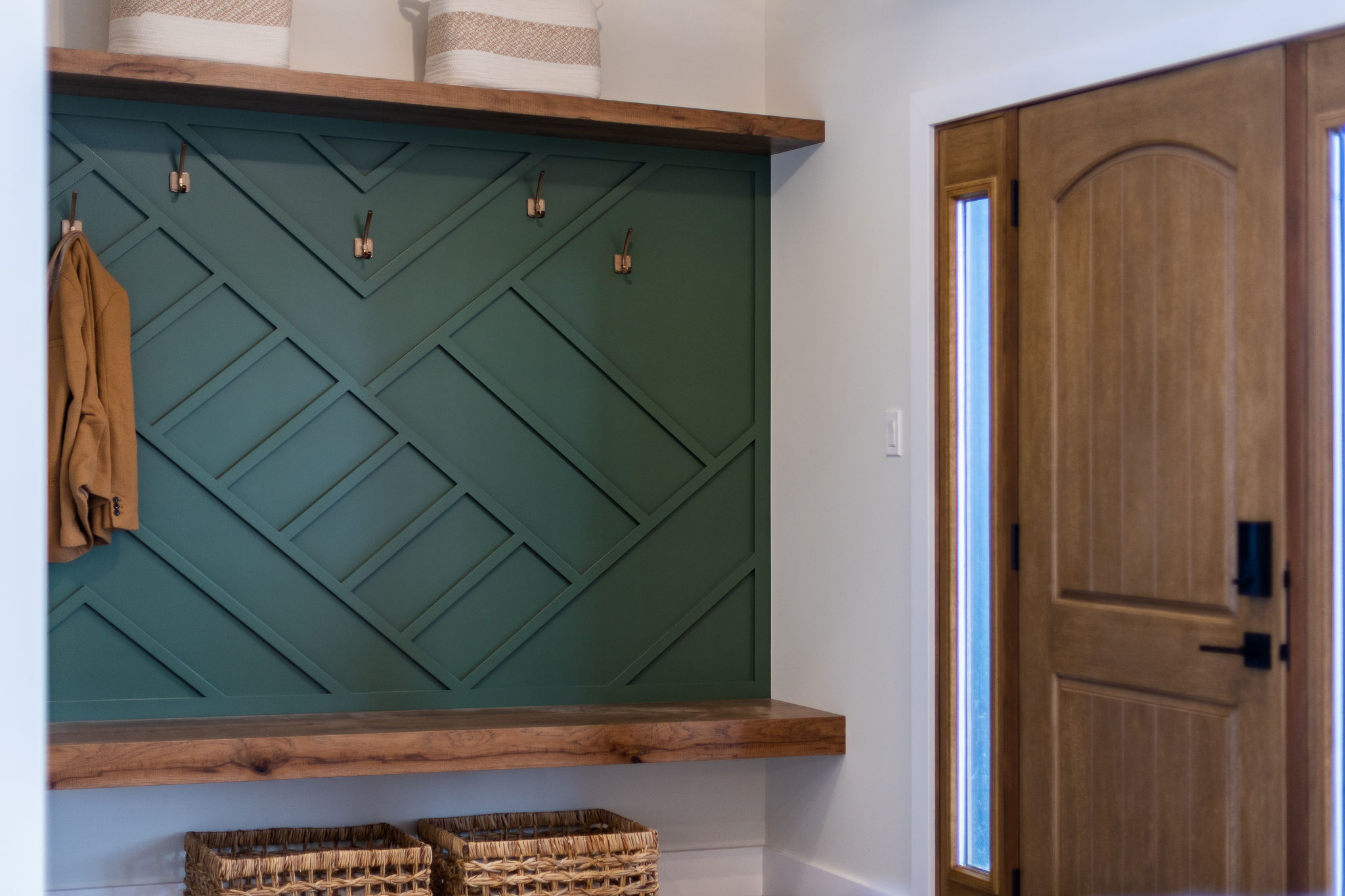 Mudroom with a green geometric accent wall, wooden bench, coat hooks with a hanging jacket, woven baskets below, and a wooden door on the right.