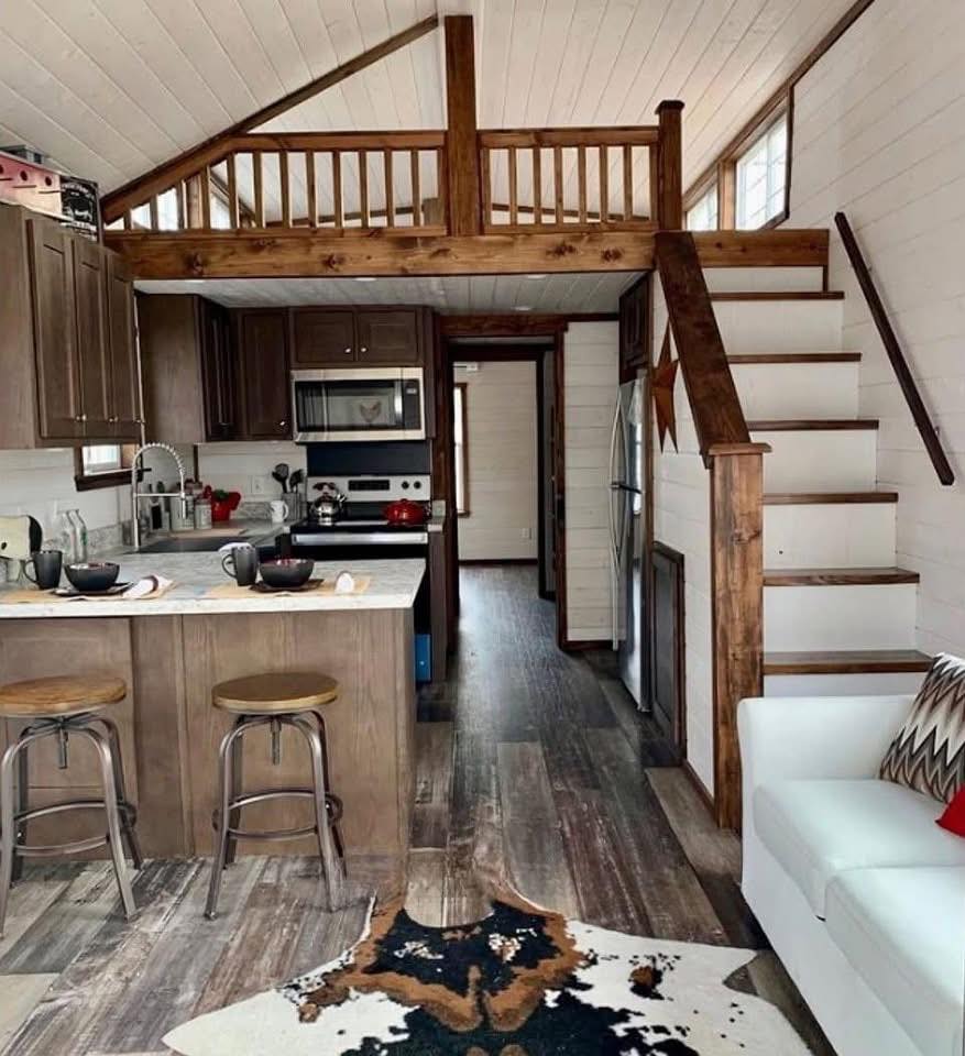 Cozy tiny house interior with a wooden loft above a compact kitchen. Features include a white sofa, rustic stools, and a cowhide rug on wooden floors.