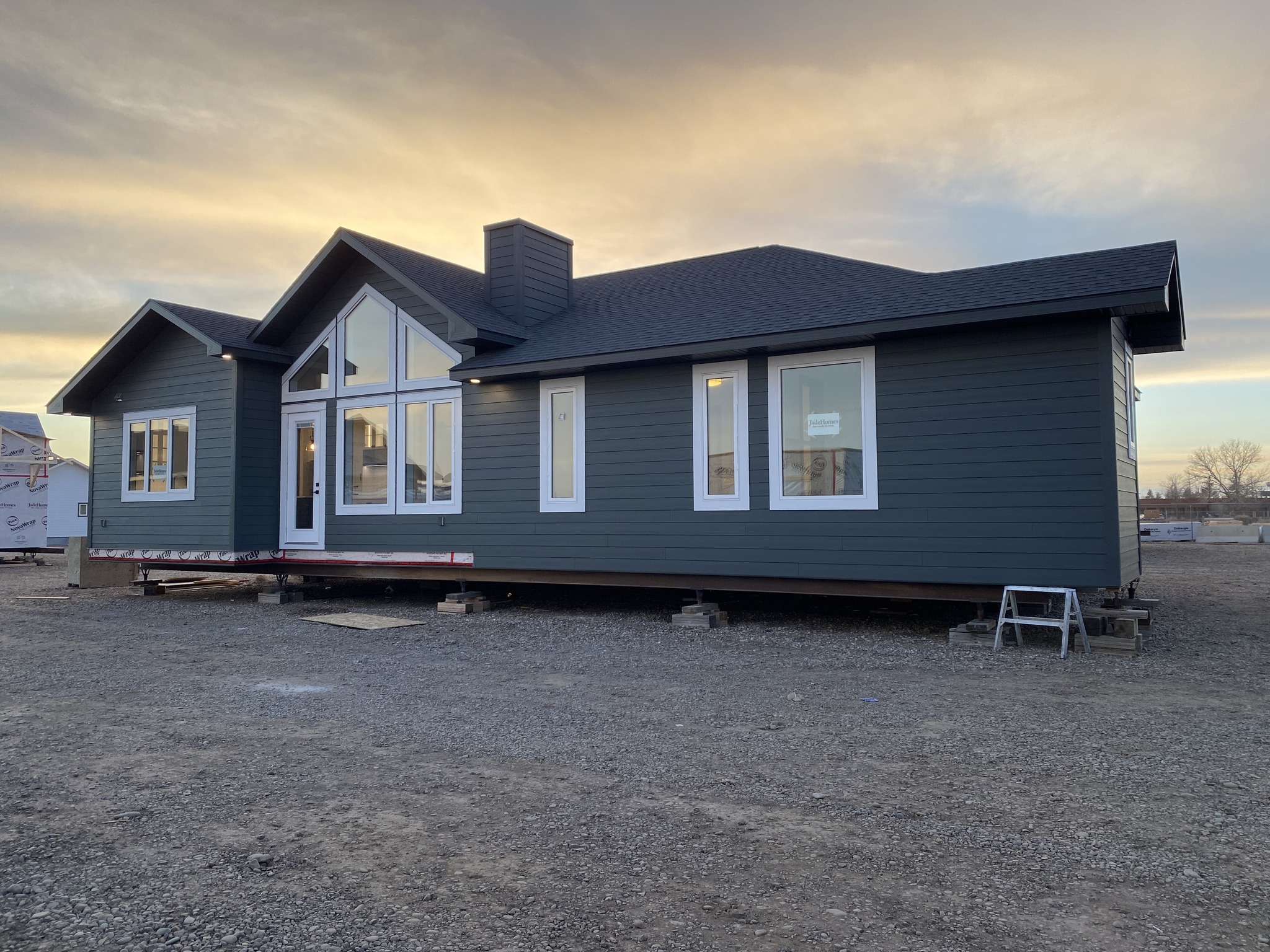 Single-story dark gray modular home with large windows and pitched roof on a gravel lot at sunset, conveying a calm and serene atmosphere.