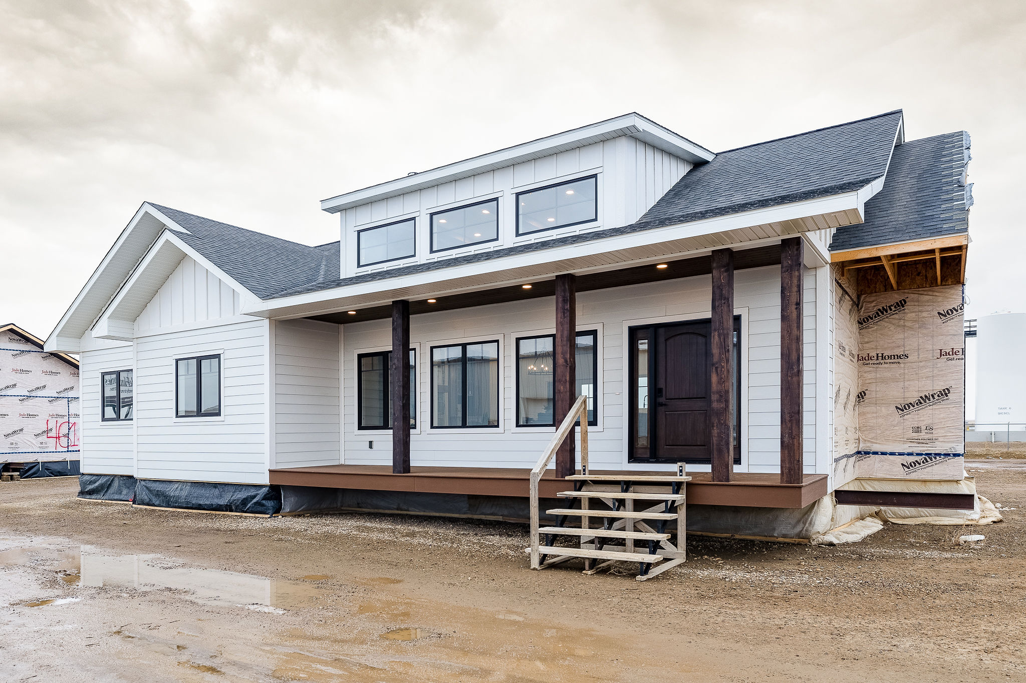 Single-story modern white house under construction, featuring a dark front door, large windows, and wooden columns. Overcast sky with a muddy foreground.