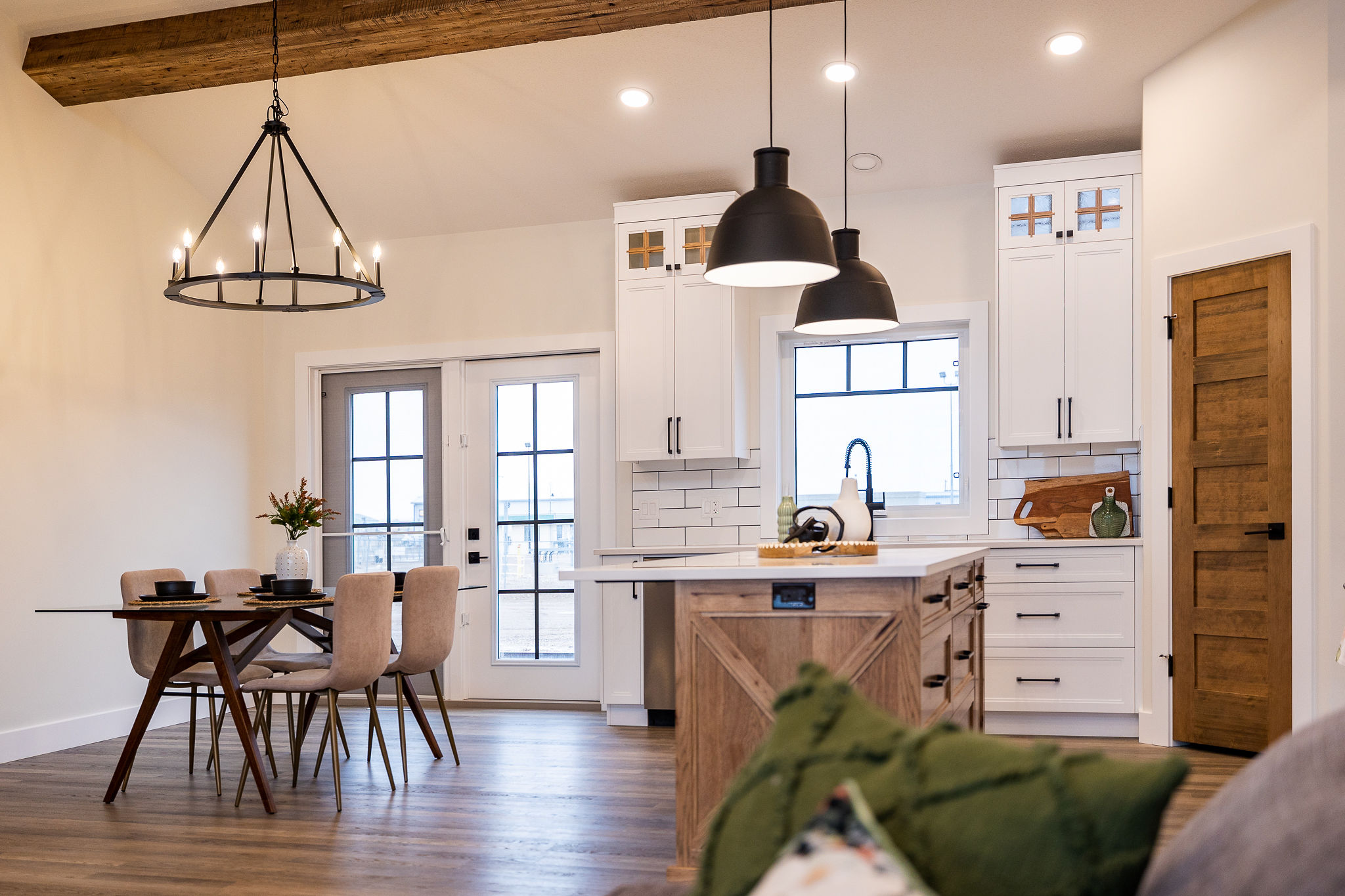 A cozy, modern kitchen with white cabinets, a wooden island, and black pendant lights. A dining table with beige chairs sits under a candle-style chandelier. Warm, inviting ambiance.