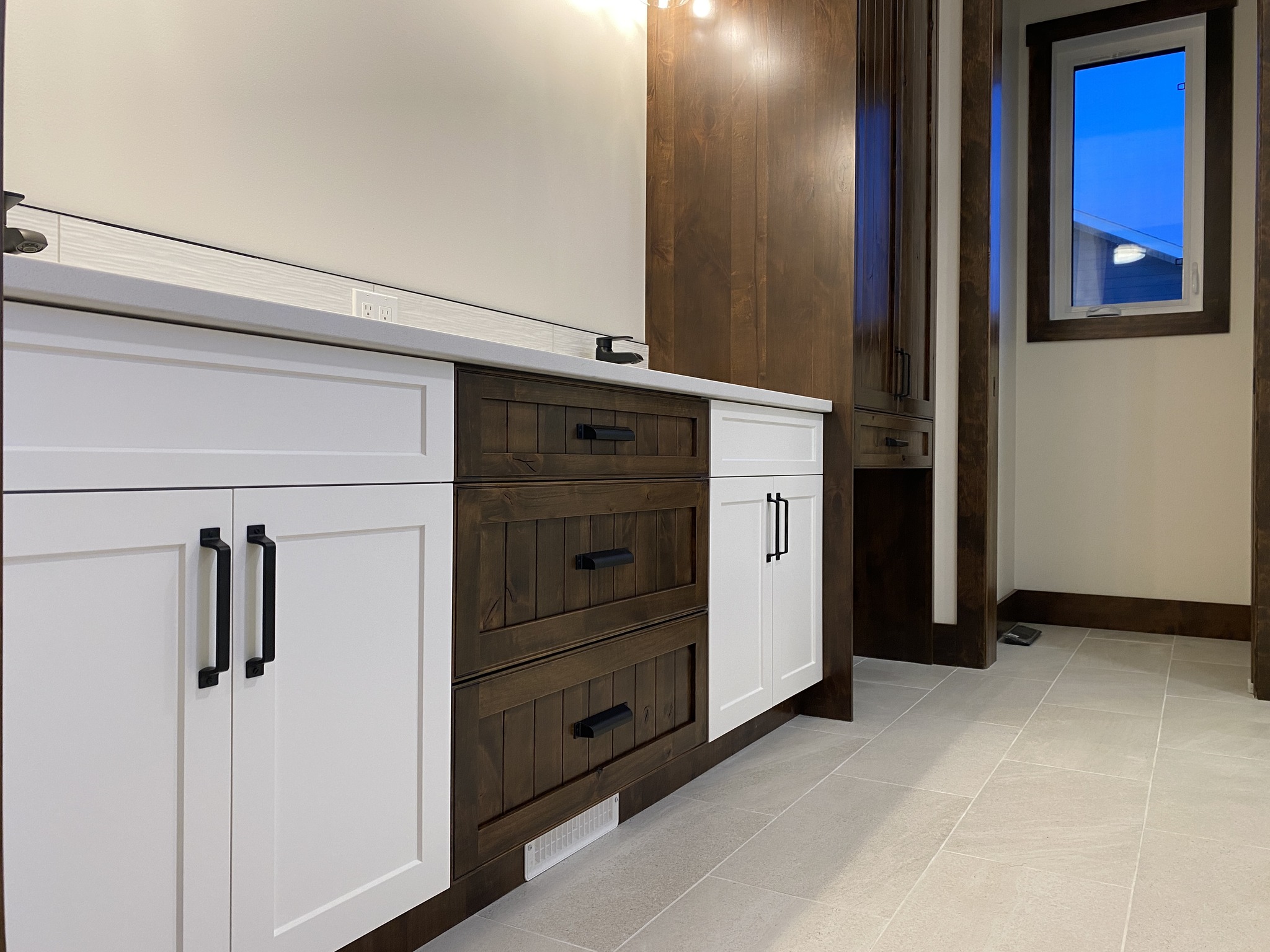 Modern bathroom with contrasting cabinets; white with black handles and dark wood with matching panels. Bright lighting and a small window enhance the space.