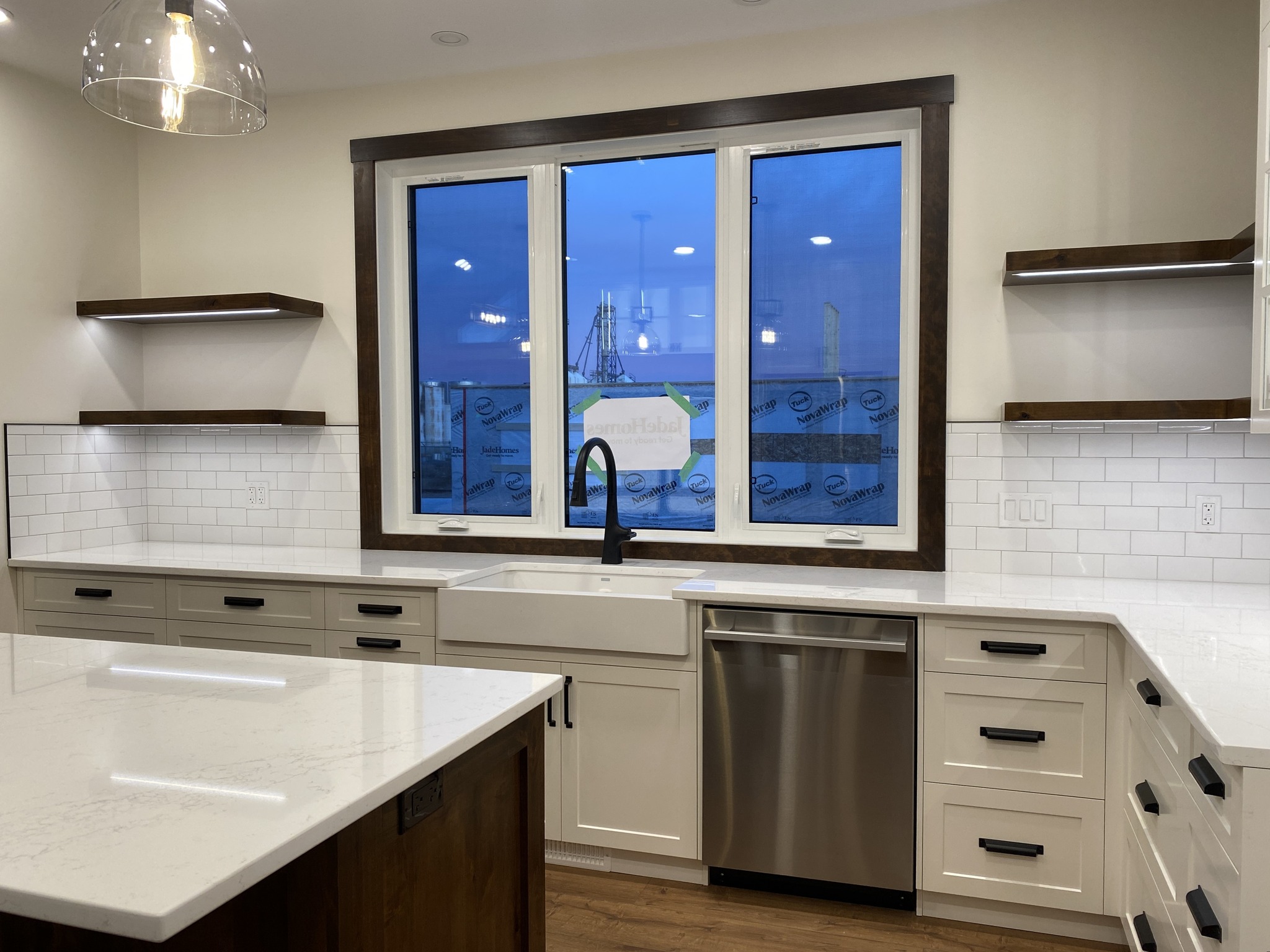 Modern kitchen with white cabinets, dark handles, and wood accents. Large central window overlooks a blue evening sky. Sleek countertops, stainless steel dishwasher, and elegant hanging light create a clean, inviting ambiance.