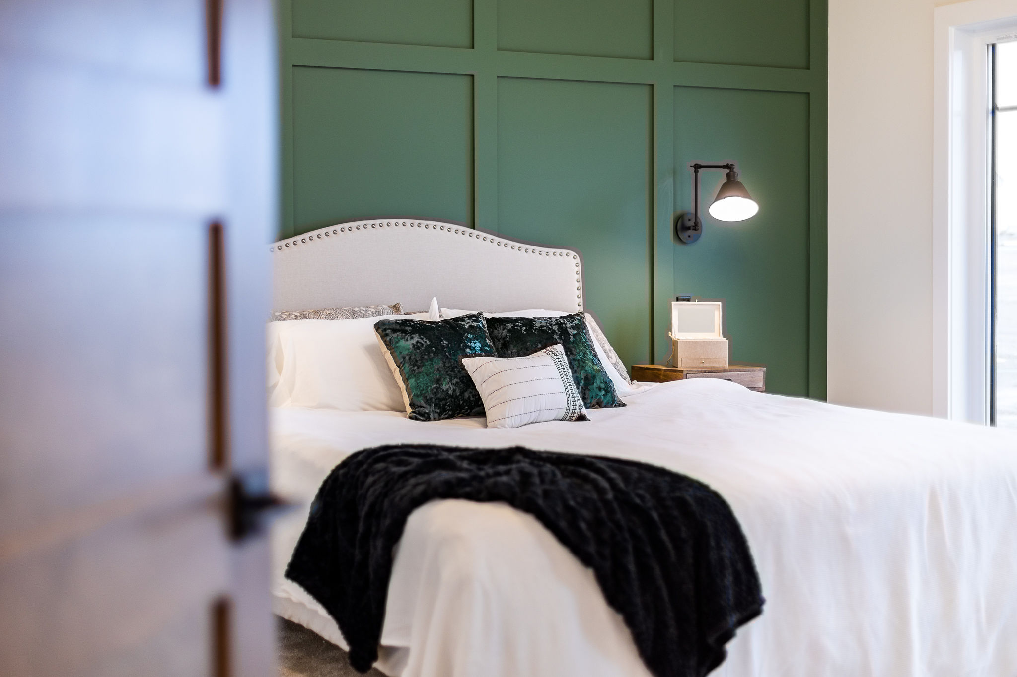 A cozy bedroom with a white, studded headboard bed, green accent wall, black blanket, and green pillows. A wall sconce provides warm lighting.