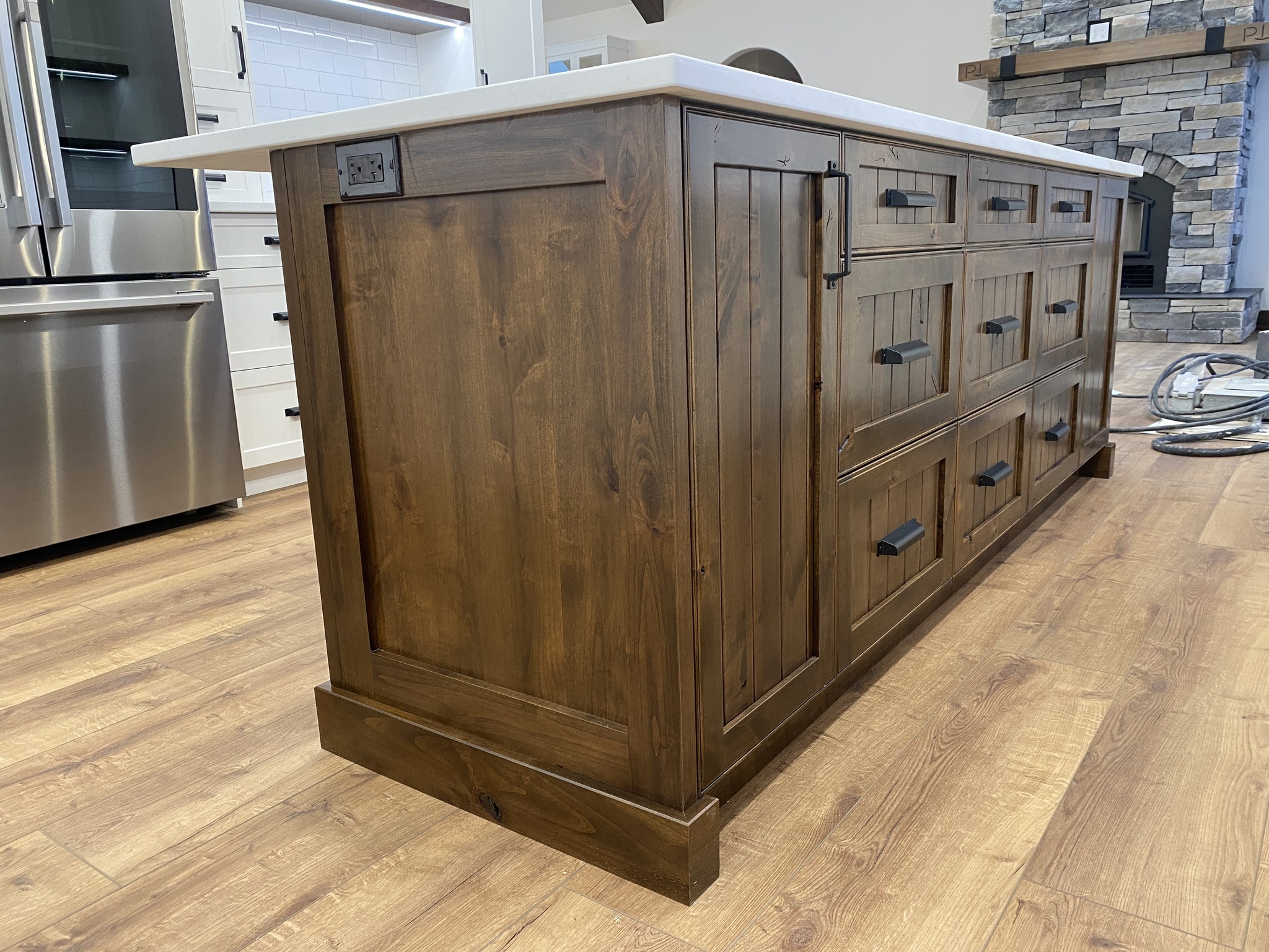 Wooden kitchen island with dark handles and a white countertop, set on a light wood floor. A stone fireplace is visible in the background.