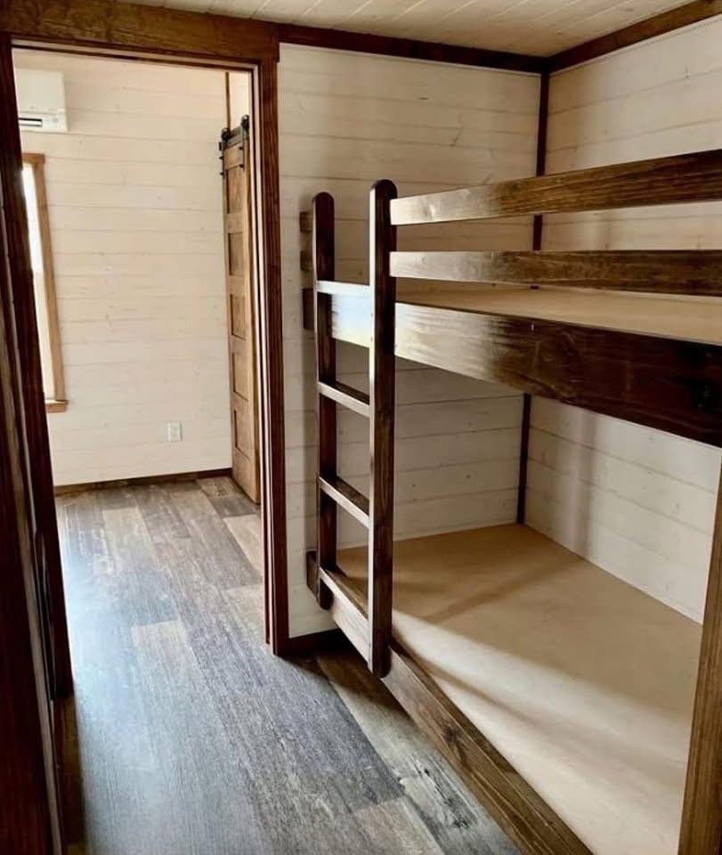 A compact room with wooden bunk beds on the right, ladder attached. Light wood walls and a rustic sliding barn door enhance the cozy, rustic charm.