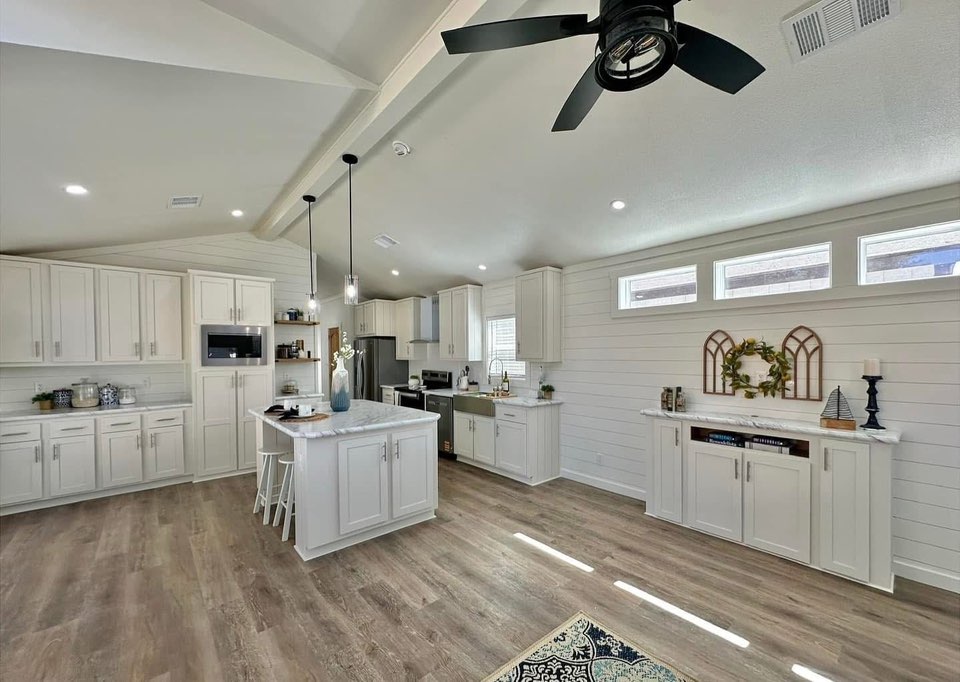 Spacious kitchen with white cabinets, island with chairs, stainless steel appliances, and light wood flooring. Bright, airy feel with modern decor and ceiling fan.