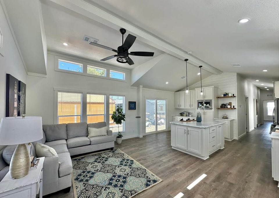 Modern open-concept living space with a white kitchen, island, and cozy gray sofa. Sunlight streams through large windows, and a ceiling fan hangs above.