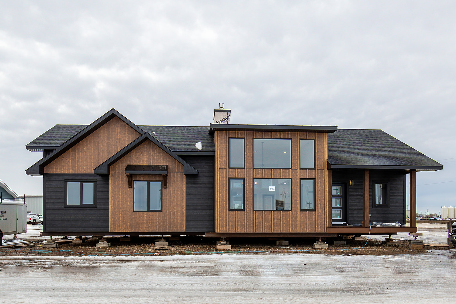 Modern single-story house with a mix of dark wood and black siding. Large windows enhance brightness. Snowy ground and overcast sky create a cold ambiance.