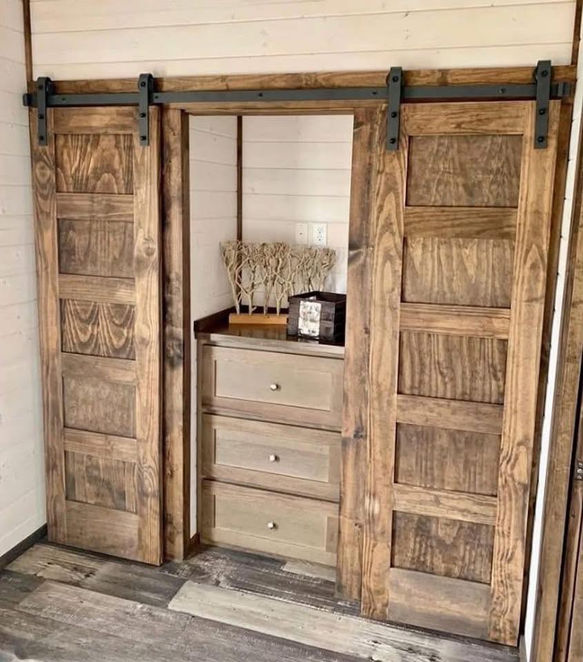 Rustic wooden sliding barn doors reveal a cozy nook with light wood drawers and decorative trees, set against white shiplap walls and gray flooring.