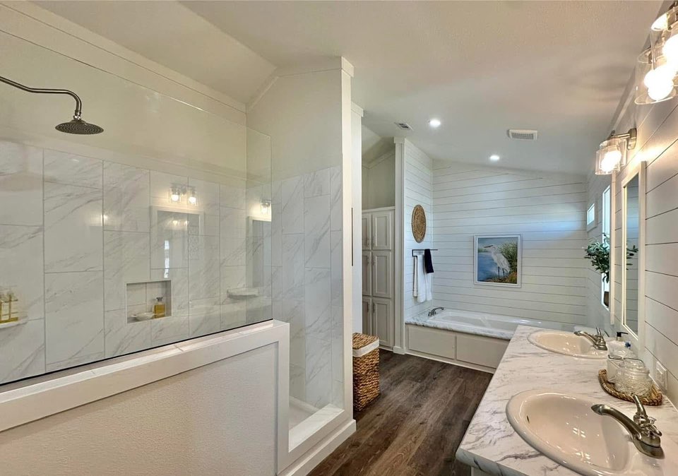 Spacious bathroom with a marble shower, freestanding tub, double sinks on a marble vanity, shiplap walls, wood flooring, and soft lighting. Calming ambiance.