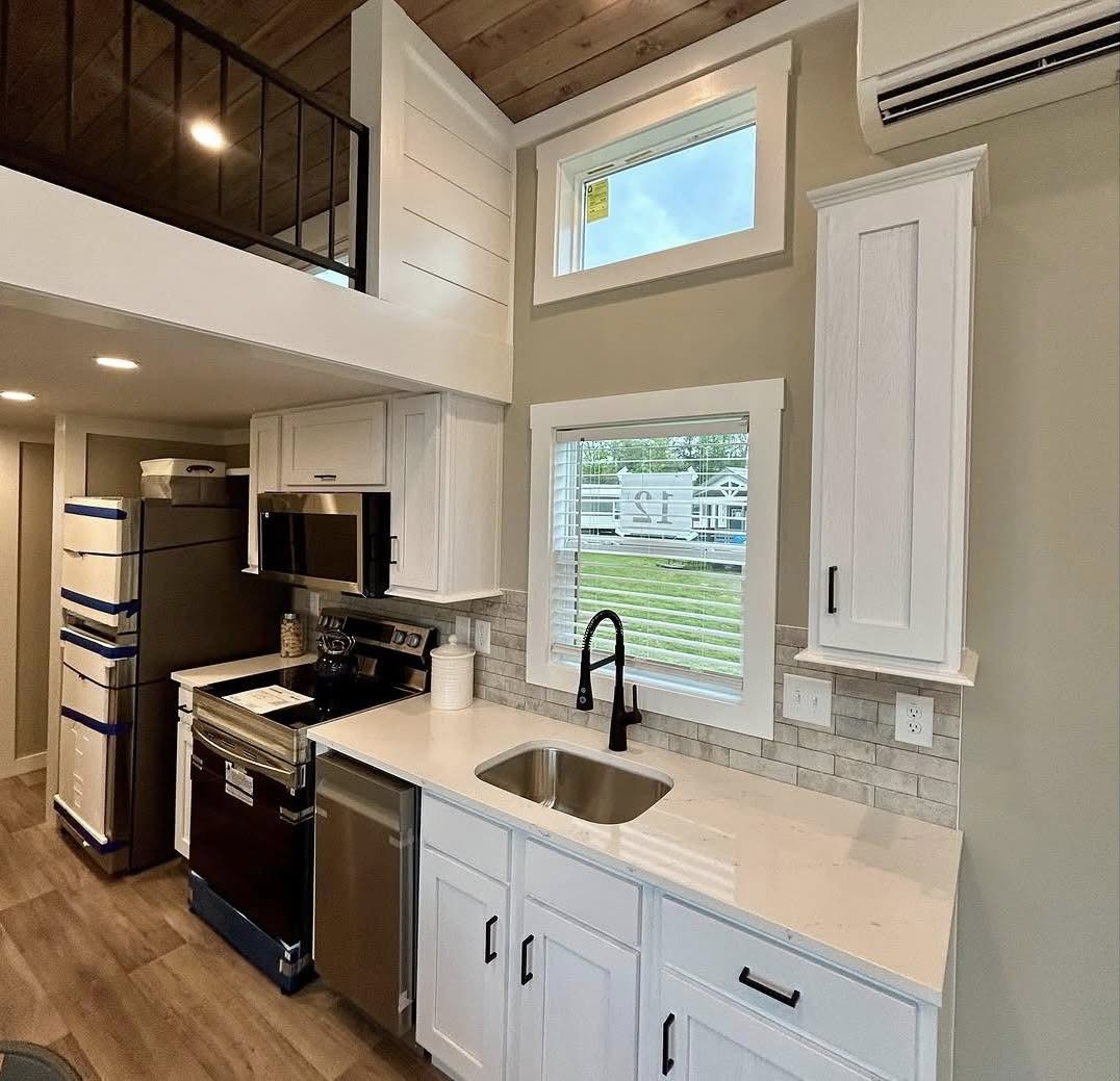 Modern tiny house kitchen with white cabinets, stainless steel appliances, and a black faucet. A loft with railing is above, creating a cozy feel.