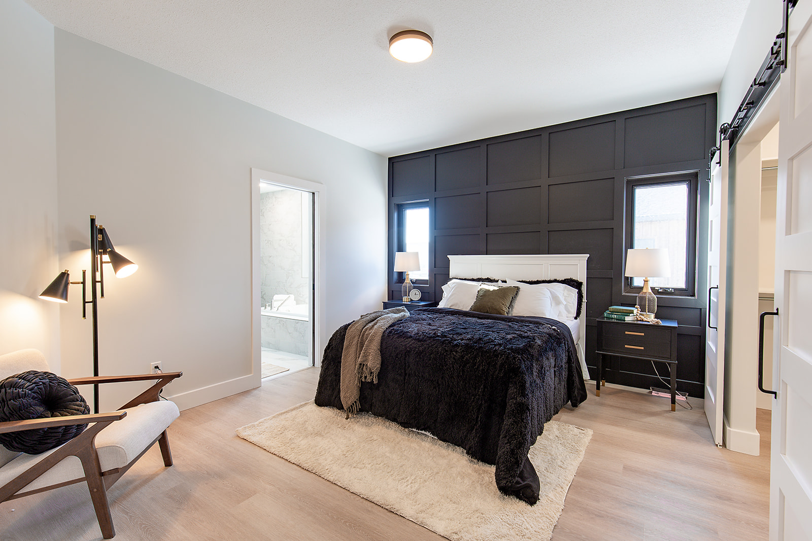 Modern bedroom with a cozy aesthetic. Features a black accent wall, plush bed, beige armchair, soft lighting, and a doorway to a sleek bathroom.