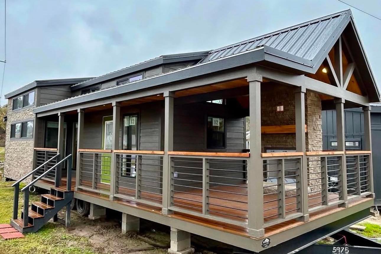 A modern tiny house with dark wood siding and a pitched metal roof. It features a wraparound porch with railing and steps, exuding a cozy, rustic vibe.