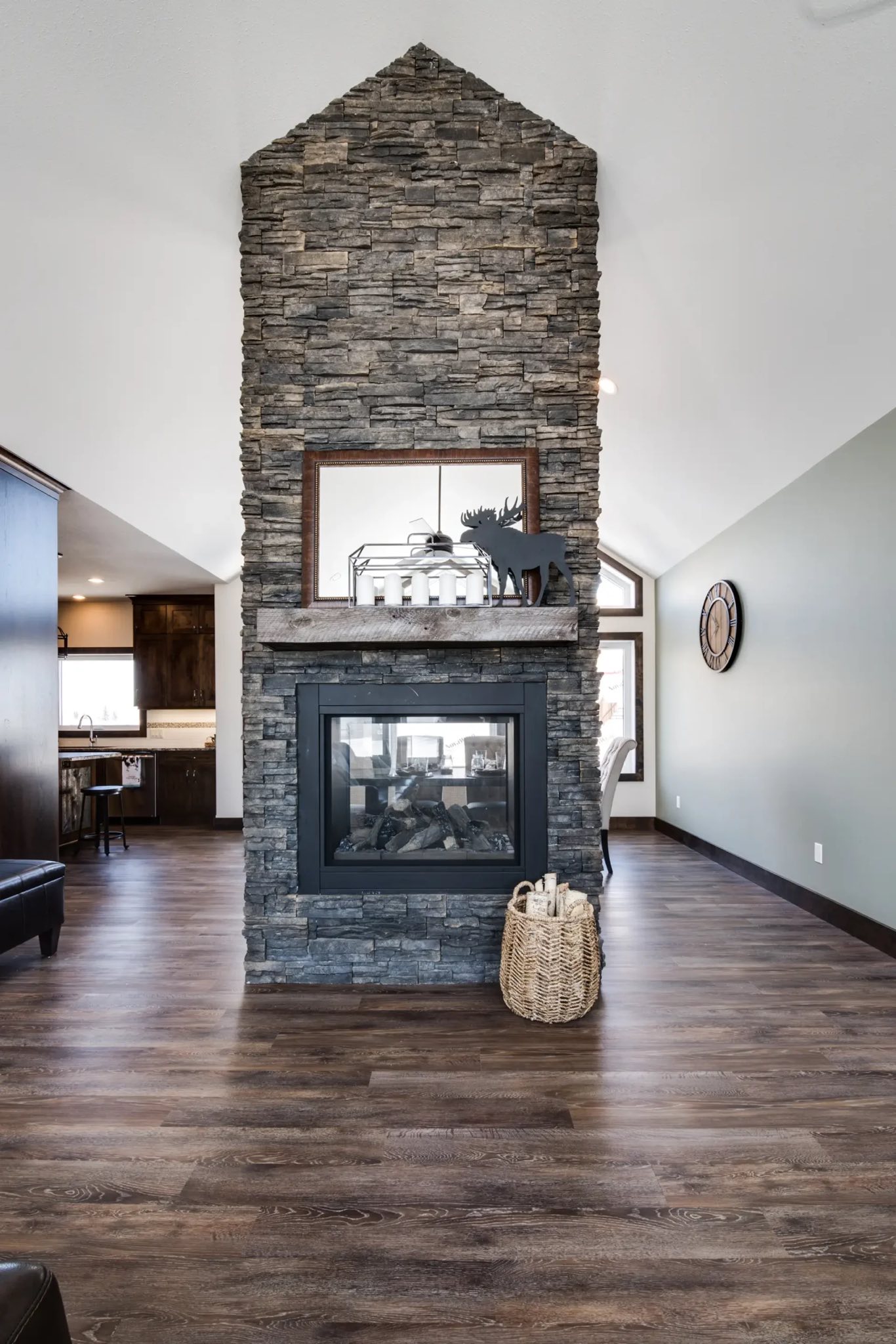 Modern living room with a tall stone fireplace and wood floors. Cozy decor includes a framed mirror, candles, and a wicker basket, creating a warm ambiance.