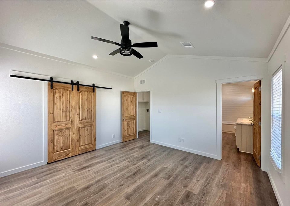 A modern, empty room featuring a light wood floor, white walls, and a black ceiling fan. Rustic wooden barn doors on the left add a warm touch.